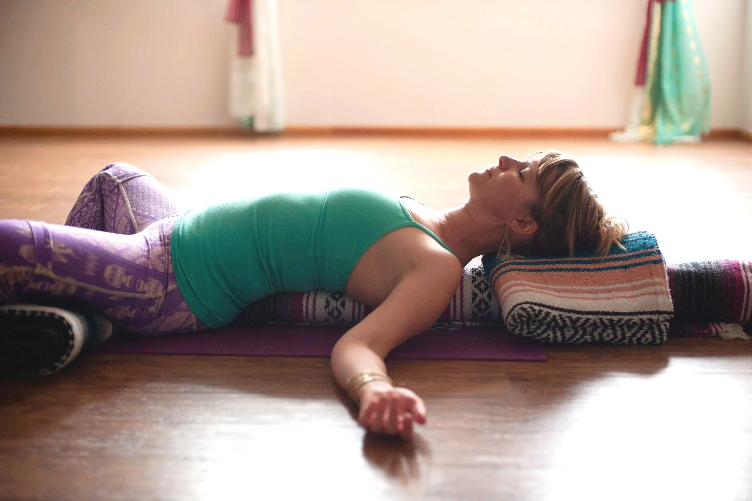 A woman lies on her back on the floor with her arm stretched out and her eyes closed, seemingly relaxing during a yoga or meditation session in a room with wooden floors and colorful curtains.