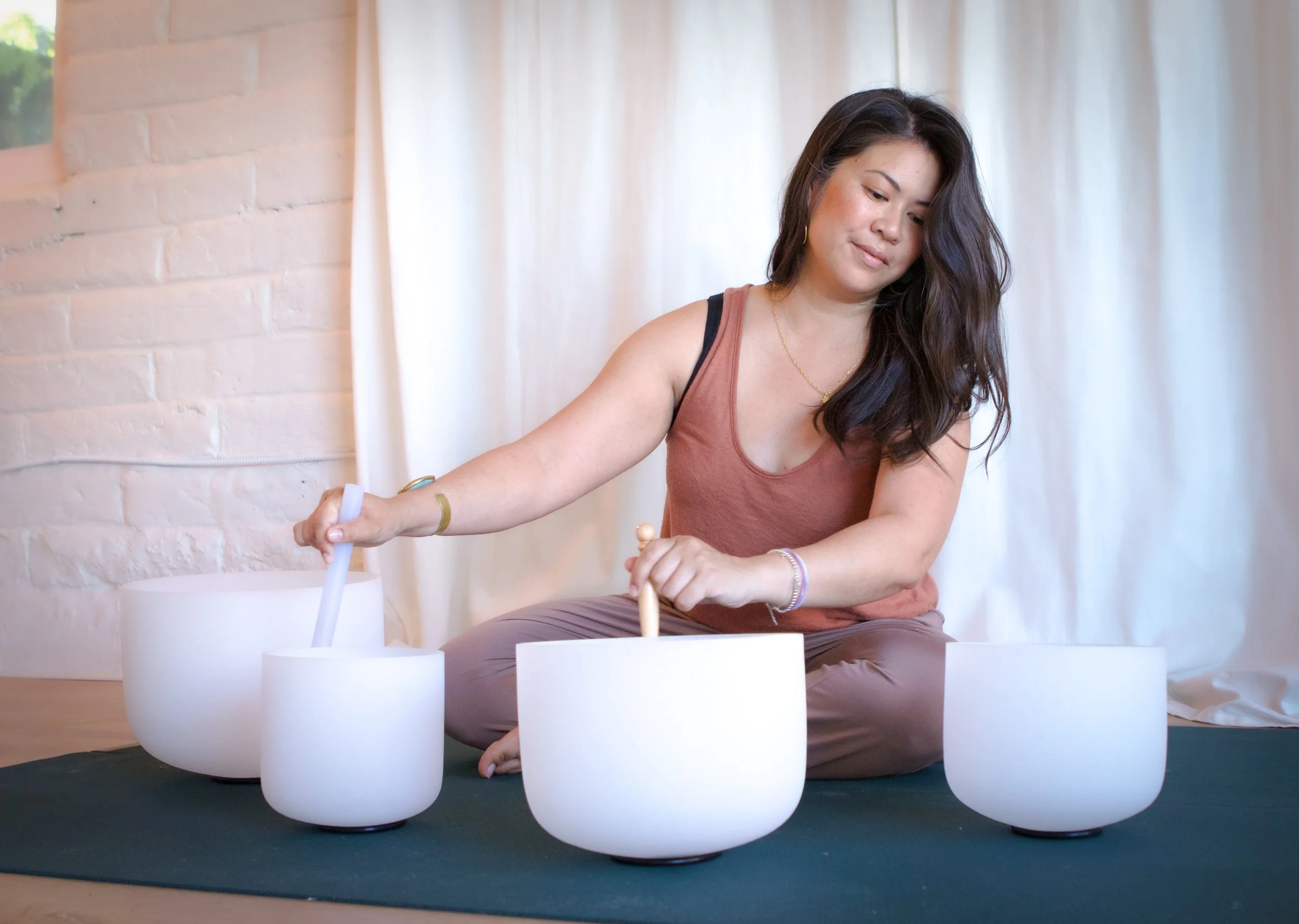 A woman with long dark hair, wearing a sleeveless top, sitting on the floor and playing a musical instrument surrounded by white drums or bowls. She is indoors with curtains and a brick wall in the background.