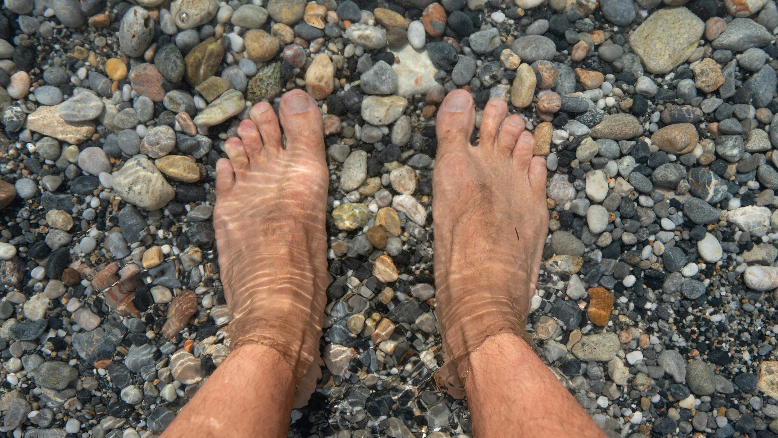 A pair of tanned feet submerged in shallow water on a pebble beach, with small waves creating ripples on the skin.