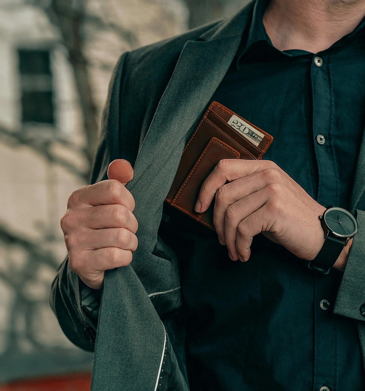 A man wearing a gray suit jacket and black shirt holding a brown leather wallet and twenty-dollar bill close to his chest in front of a blurred building background.