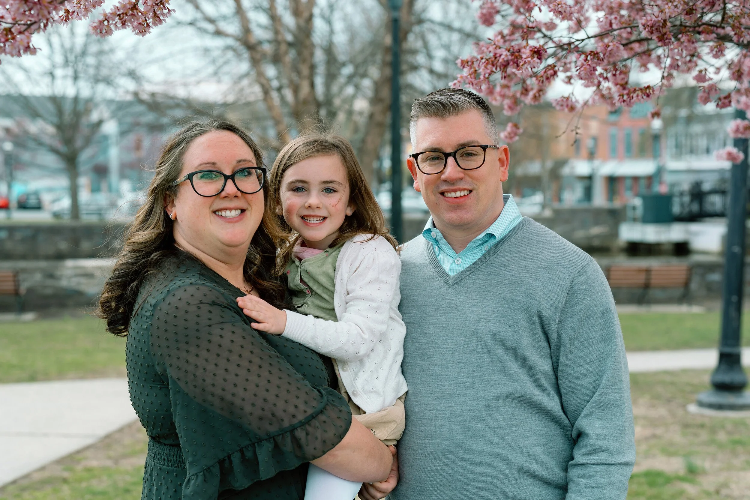 A family of three smiling outdoors in front of pink cherry blossom trees, with houses and a park in the background.