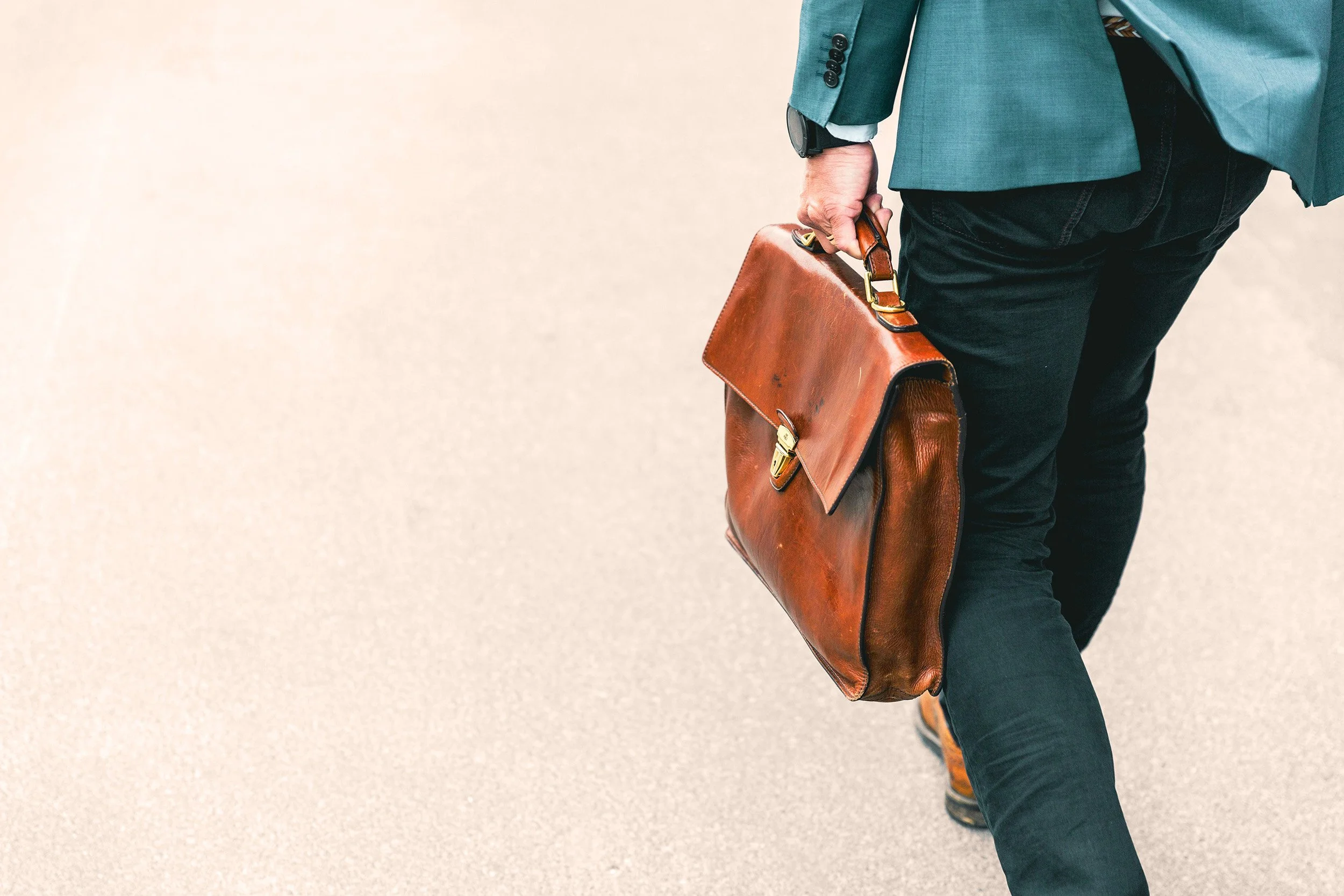 A man in dark pants and a teal blazer walking on a beige sidewalk, carrying a worn brown leather briefcase.