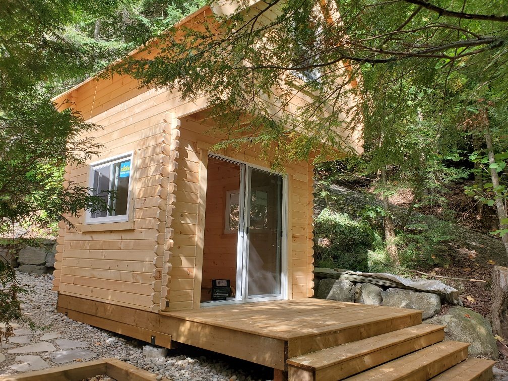Small wooden cabin surrounded by trees in a forested area, with a wooden deck and glass doors.