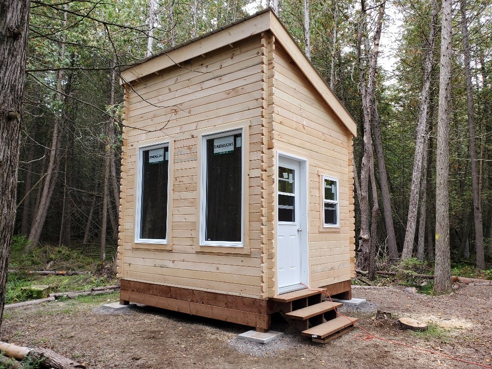 A small wooden cabin in a forest setting with large windows and a white door. It has a sloped roof and is elevated on blocks with a few steps leading up to the entrance.