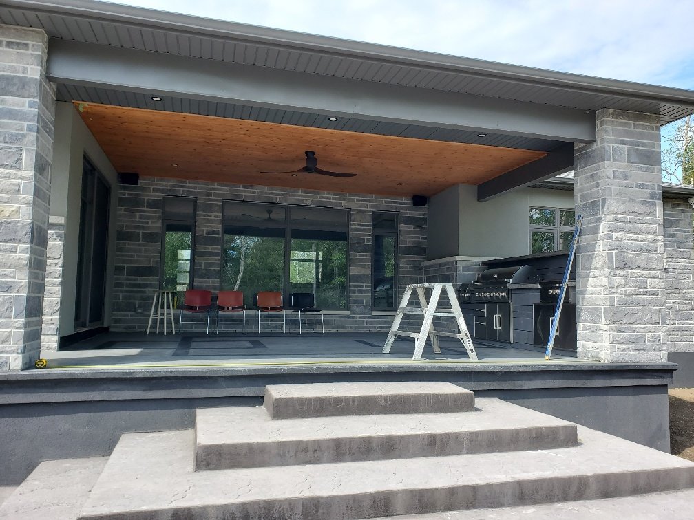 Modern patio area with gray stone walls, steps leading up, outdoor furniture, and a grill setup. Includes a wooden ceiling with a ceiling fan and a ladder leaning against the wall.