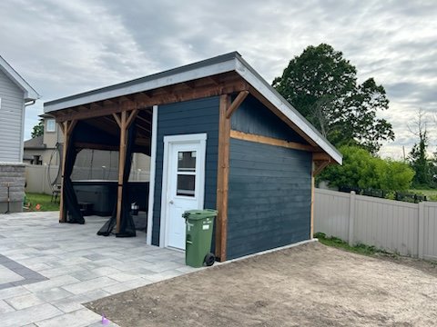 Small wooden shed with white door, located in a backyard near a paved area and fenced surroundings.
