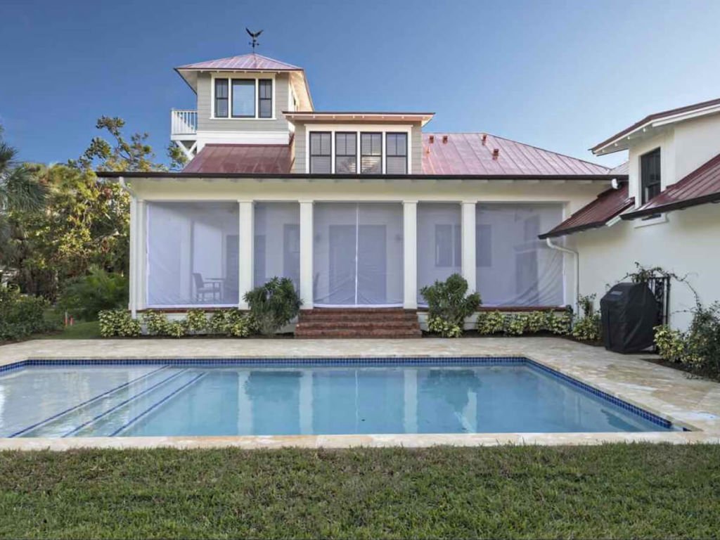 Colonial-style house with a red metal roof, white columns, and screened porch, facing a rectangular swimming pool and surrounded by greenery.