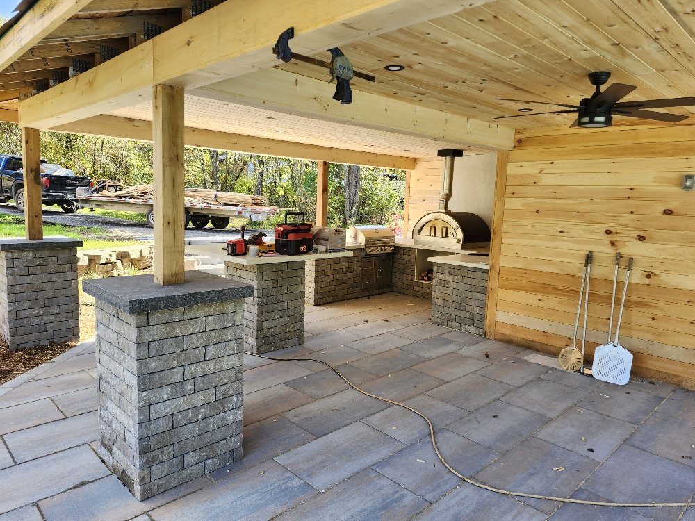 Outdoor kitchen under a wooden pergola with stone counters, pizza oven, grill, tools, and a ceiling fan.