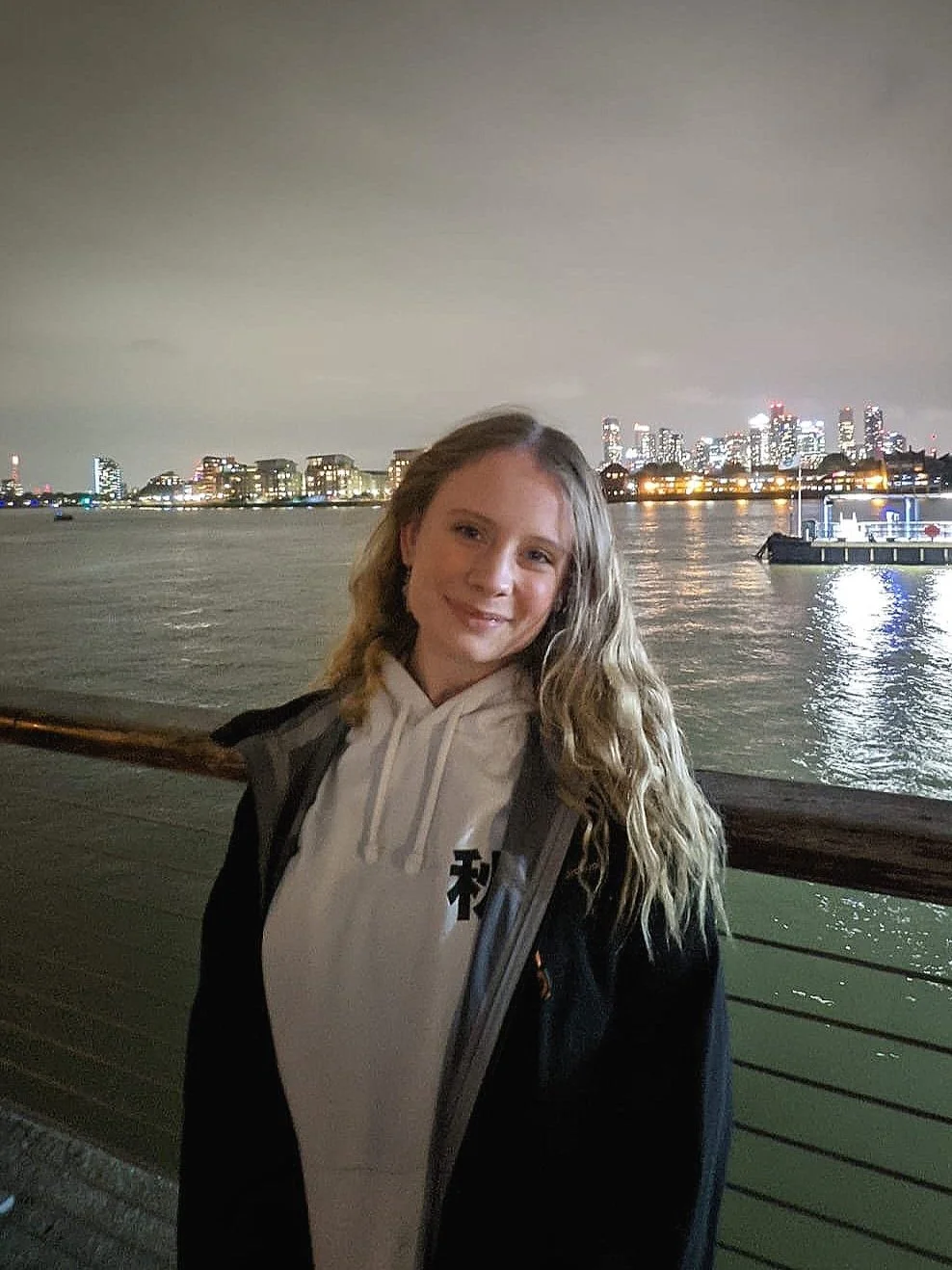 Young woman with long blonde hair smiling by a waterfront at night, city skyline with illuminated buildings in the background.