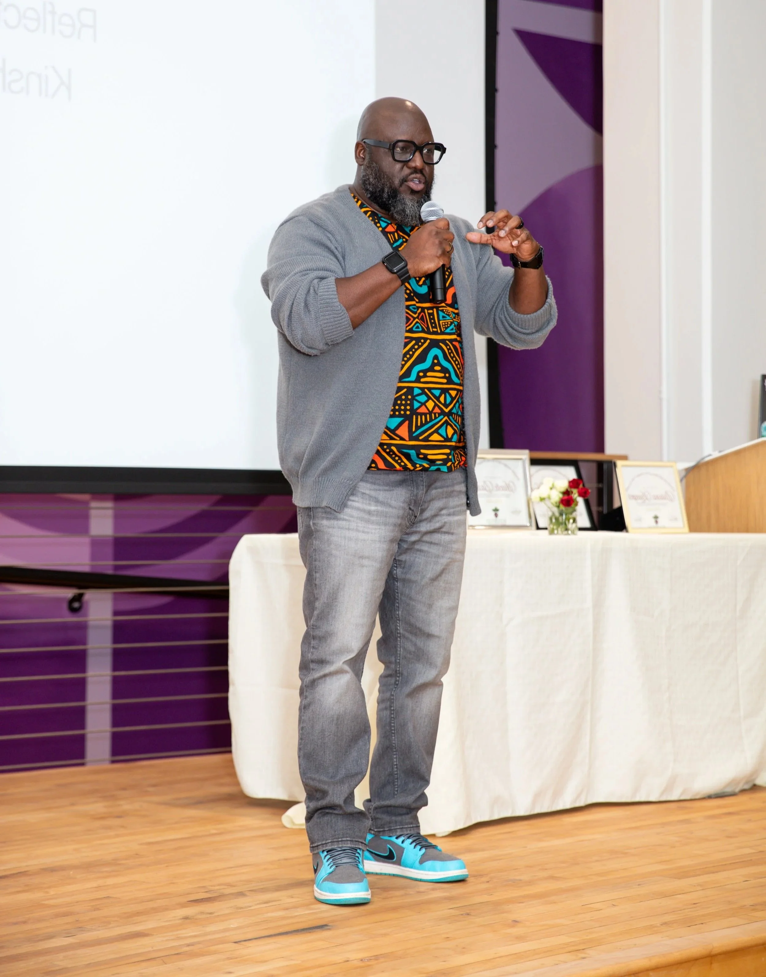 A man with glasses and a gray beard, wearing a colorful African-patterned shirt, a gray cardigan, jeans, and blue sneakers, is speaking into a microphone in front of a white projection screen at an indoor event.