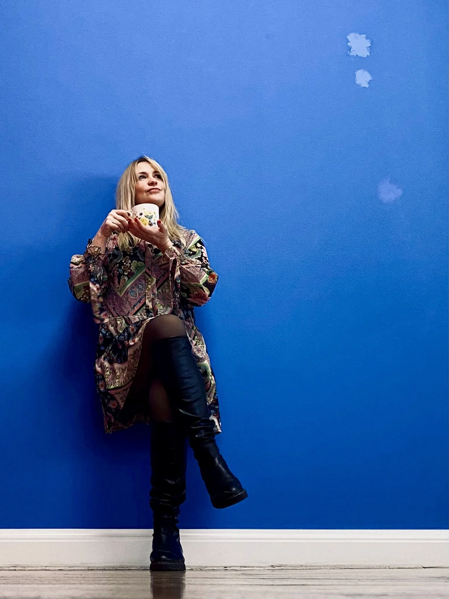Junge Frau in buntem Kleid mit hohen schwarzen Stiefeln lehnt gegen eine blaue Wand und hält eine Tasse, Blick nach oben.