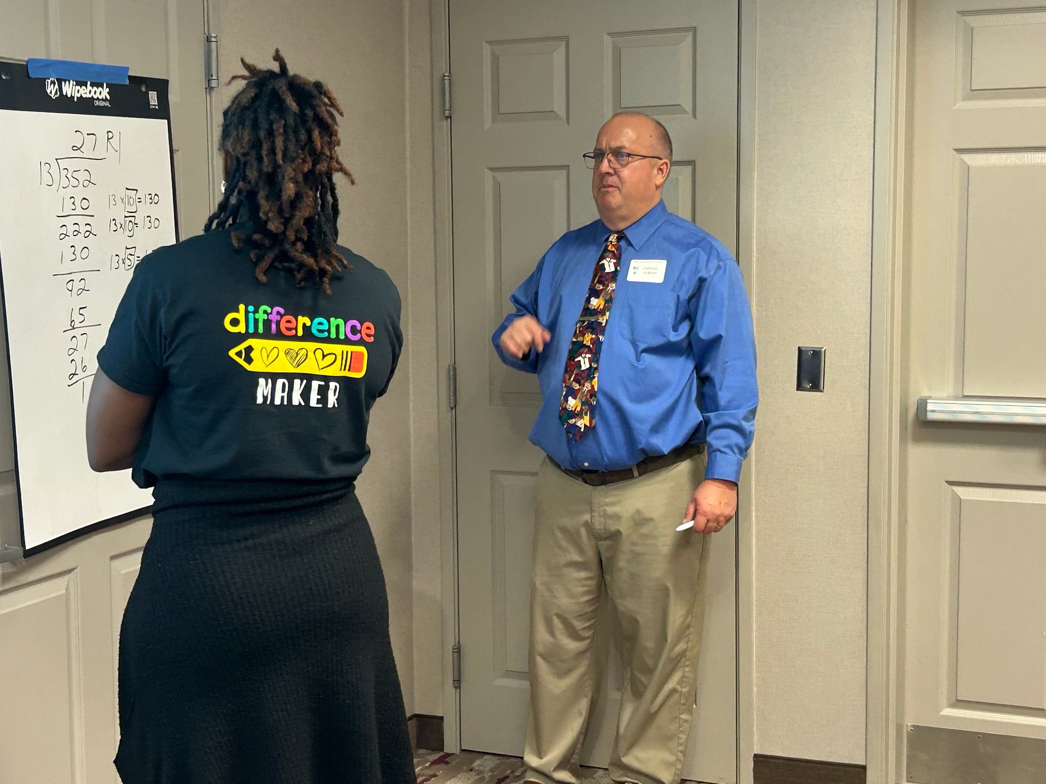 A woman and a man are having a conversation in an indoor setting, with a whiteboard visible on the left side of the image, and a beige door behind the man. The woman has dreadlocks and wears a black shirt with colorful text, and the man is dressed in a blue shirt with an advanced tie pattern and glasses.
