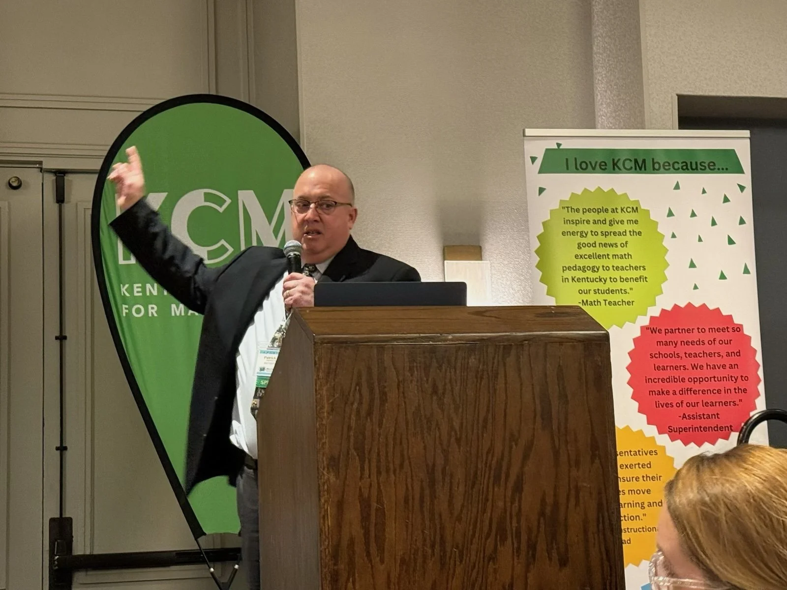 A man in a suit and glasses giving a presentation at a conference, standing behind a wooden podium, with a microphone in hand, gesturing with his left hand, and standing next to a green and white banner that reads 'KCM Kentucky for M' and a colorful poster with quotes.