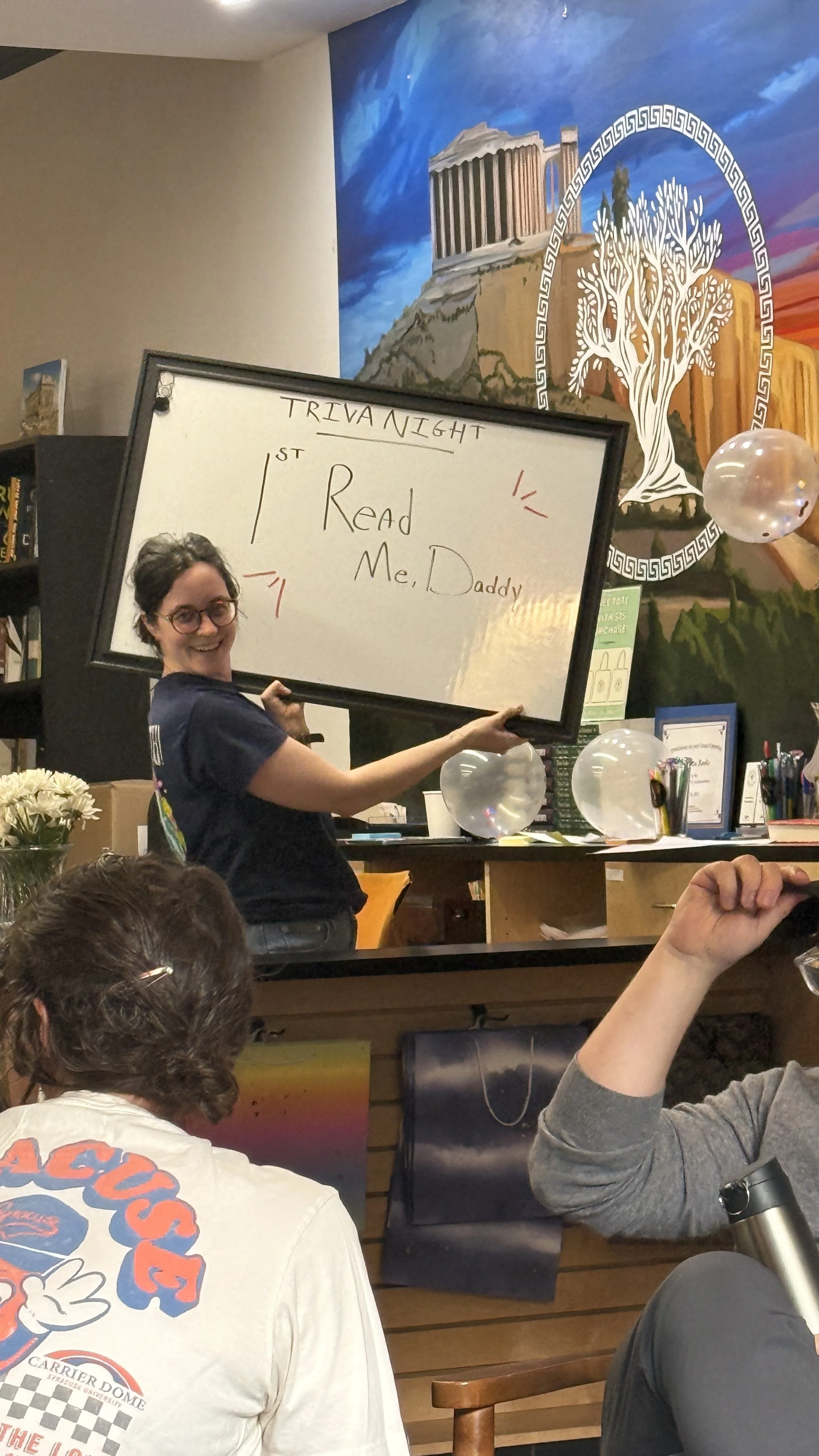 A woman holding a whiteboard with the handwritten words 'TRIVIA NIGHT, 1st, Read Me, Daddy' in a lively indoor setting with a colorful mural of ancient Greek architecture and white balloons.