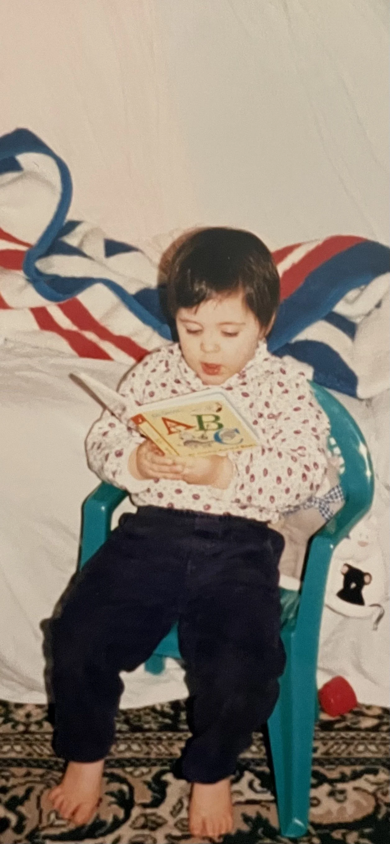 A young child with short dark hair sitting in a teal plastic chair, reading a colorful children's book, wearing a white shirt with red and purple dots and dark pants, inside a room with a patterned rug and a bed with a striped blanket in the background.