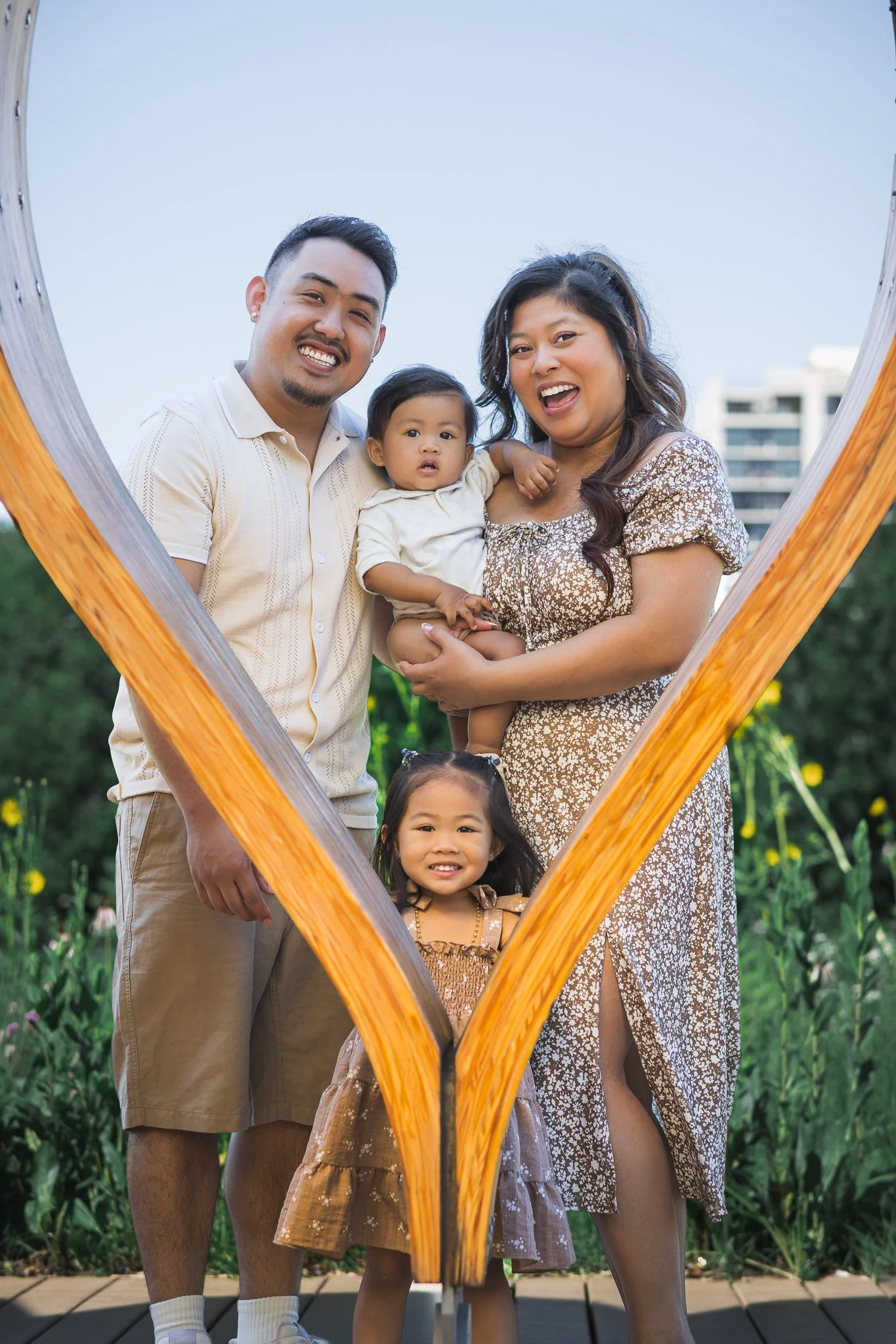 A smiling family of four posing outdoors, framed by a wooden heart-shaped structure, in a garden with lush green plants and a building in the background.