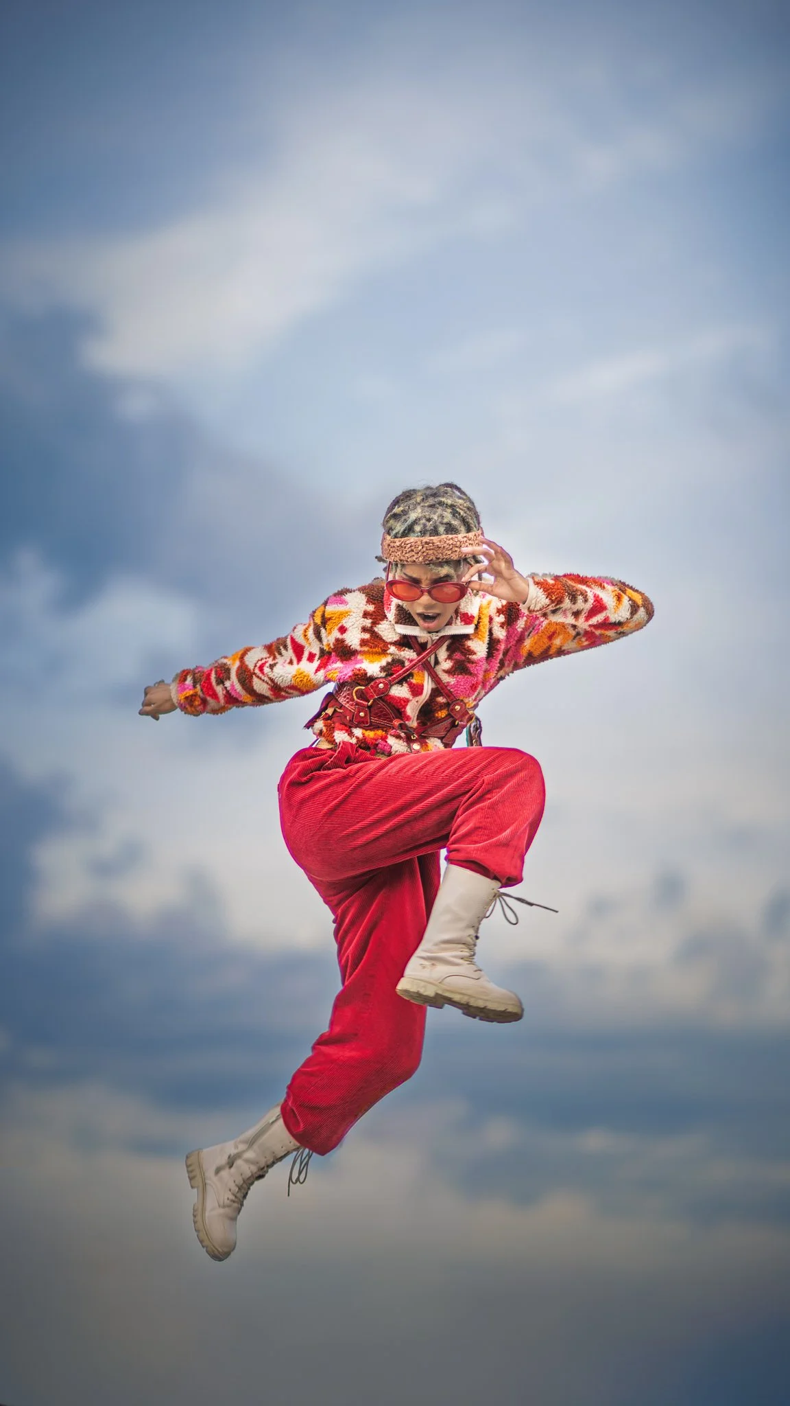 A person dressed in vibrant, colorful 80s-inspired fashion, including red pants, a patterned sweater, and boots, appears to be mid-air with one leg up and an expressive facial expression, set against a cloudy sky.