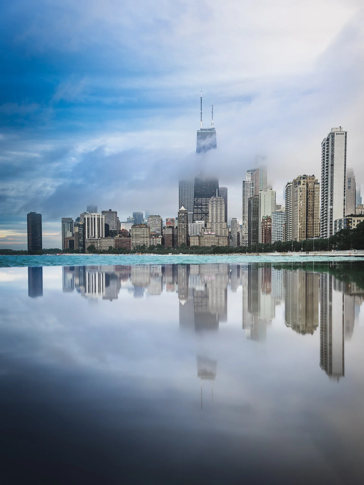 Skyline of a city with high-rise buildings partially covered by fog, reflected in a body of water in the foreground.