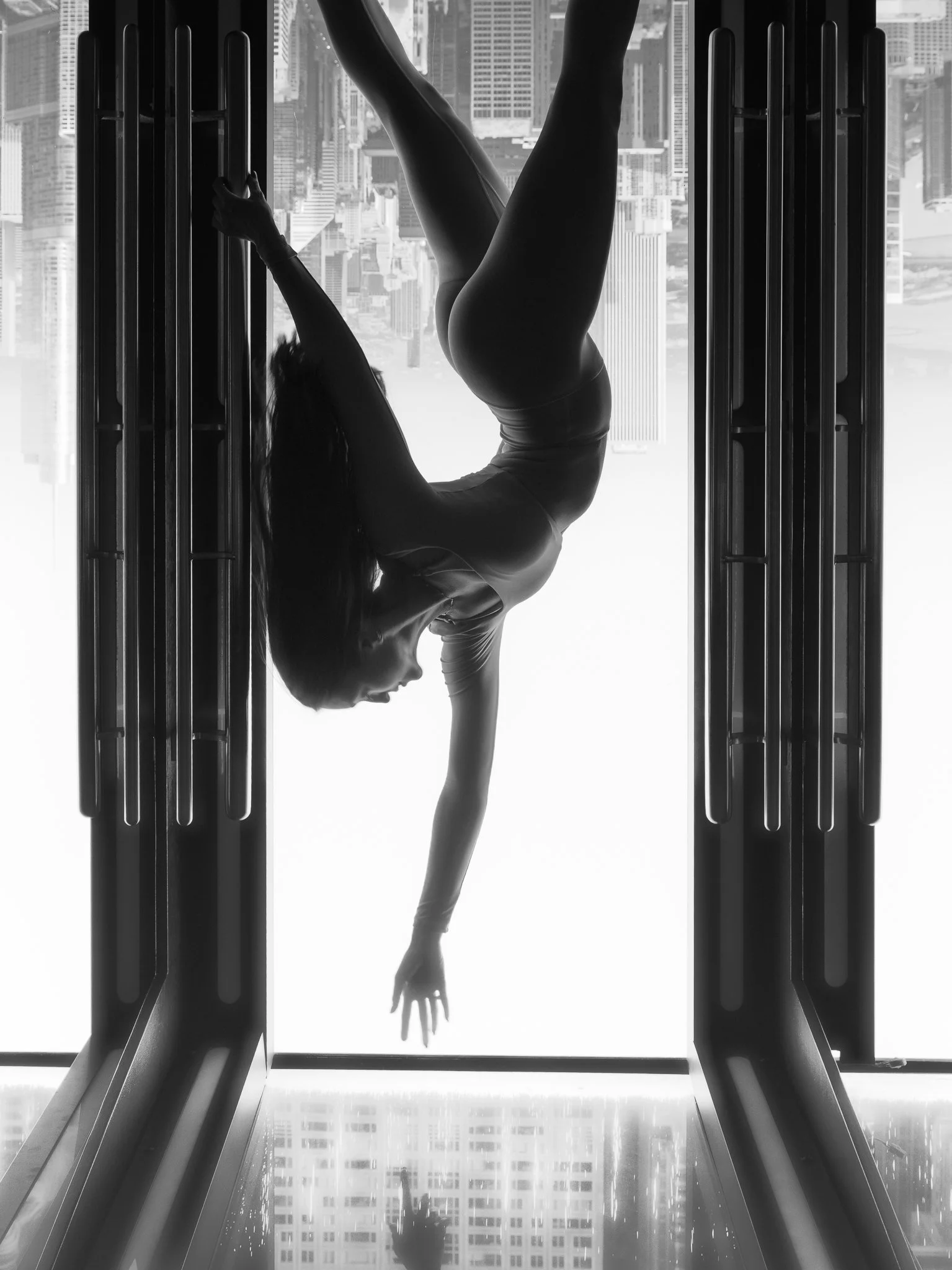A person performing a handstand inside an escalator shaft in a city skyline with tall buildings, viewed from below in black and white.
