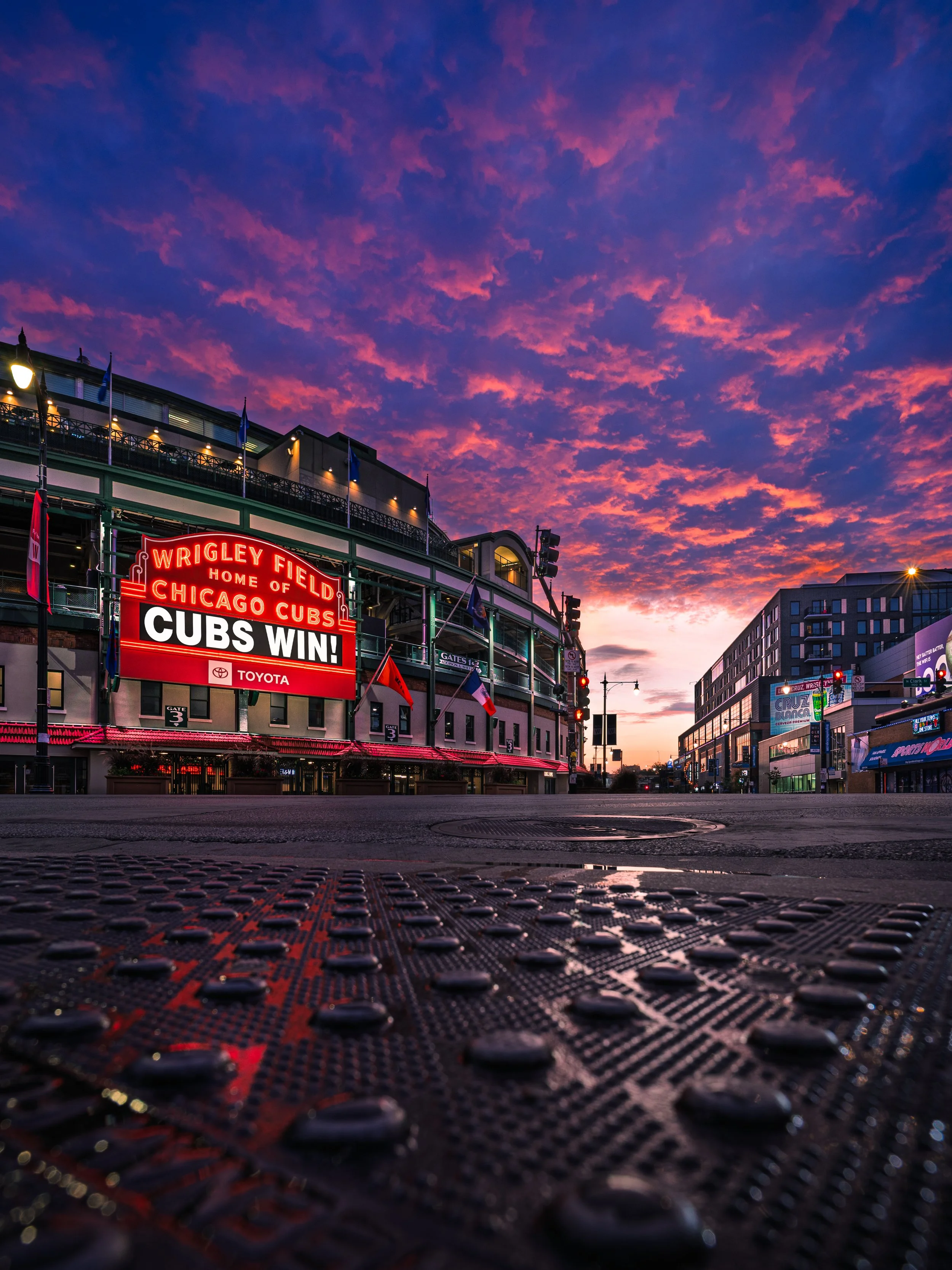 Empty street outside Wrigley Field baseball stadium during sunset, with colorful clouds and several flags, including the Chicago Cubs flag. A digital scoreboard displays a victory message for the Cubs.