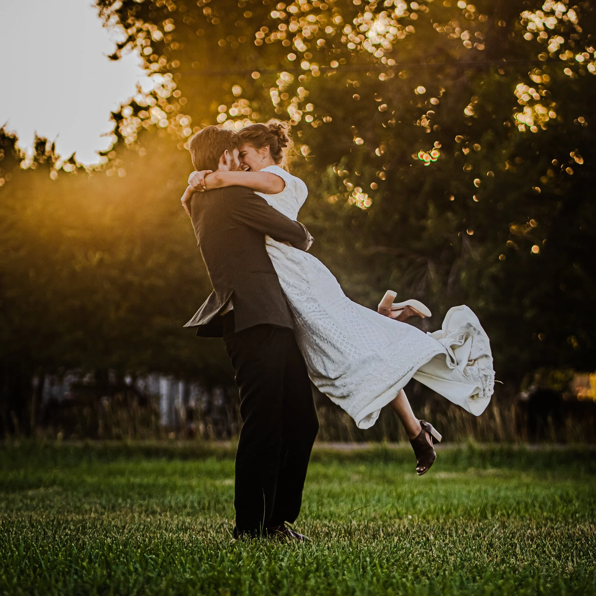A couple celebrating their wedding, with the groom lifting the bride off the ground in a field during sunset.