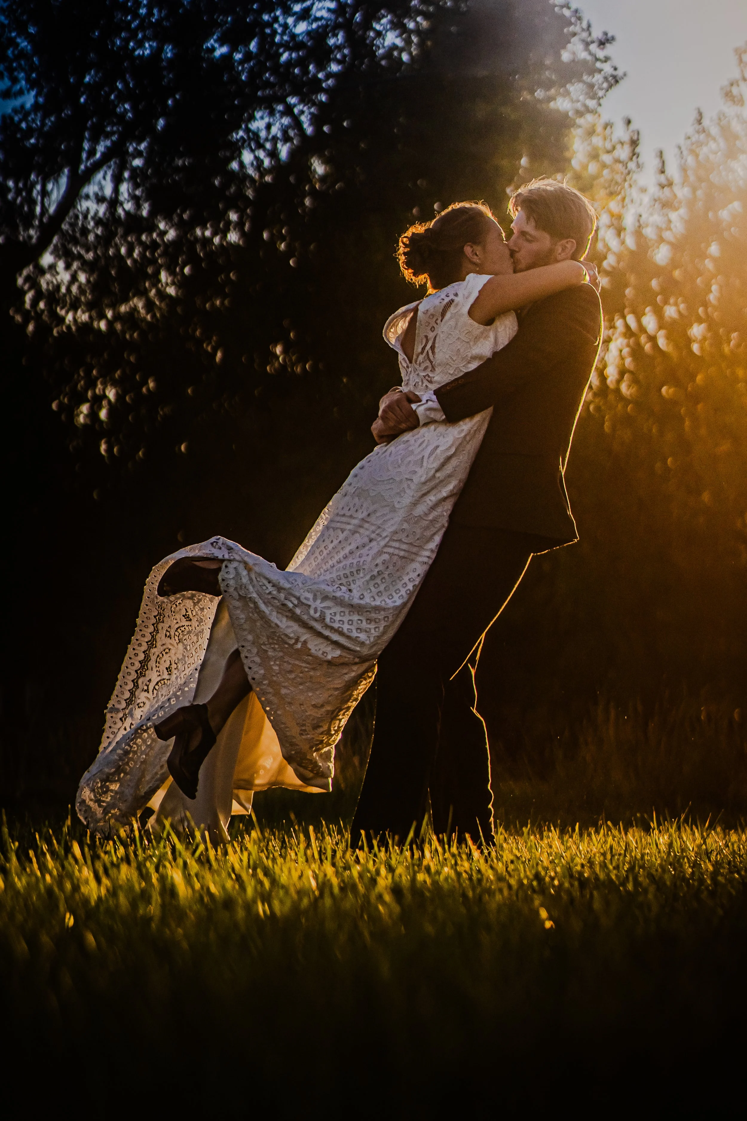 A couple, with the man in a black suit and the woman in a white lace dress, sharing a kiss outdoors at sunset, with the man lifting the woman in his arms.