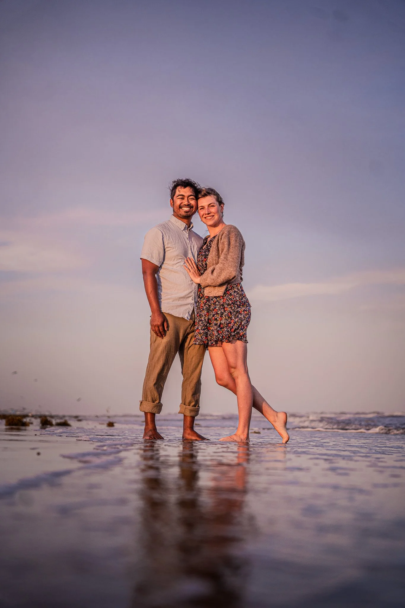 A happy couple standing arm in arm at the beach during sunset, barefoot on the wet sand with gentle waves in the background.