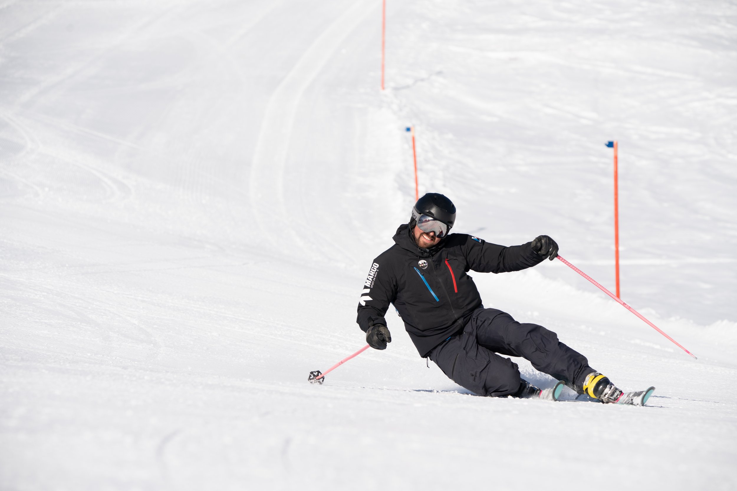 Ein Skifahrer fährt in einer sportlichen Haltung eine schneebedeckte Piste hinunter. Er trägt schwarze Skikleidung, einen Helm und eine Skibrille. Im Hintergrund sind rote Stangen zur Markierung der Strecke zu sehen.