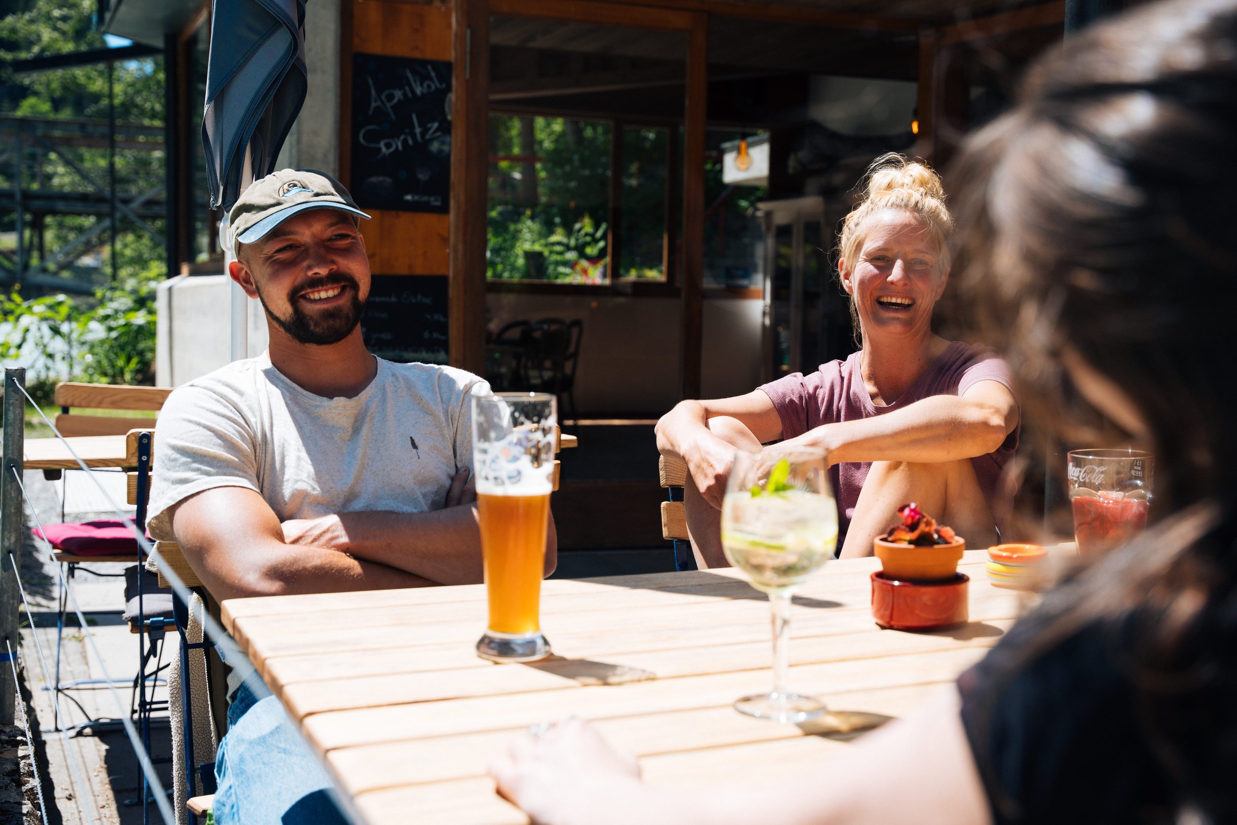 Drei Personen sitzen an einem Holztisch im Freien, lächeln und unterhalten sich. Auf dem Tisch stehen Getränke, darunter ein Glas Weißbier und ein Glas mit einem Cocktail. Die Personen scheinen entspannt und die Sonne scheint.
