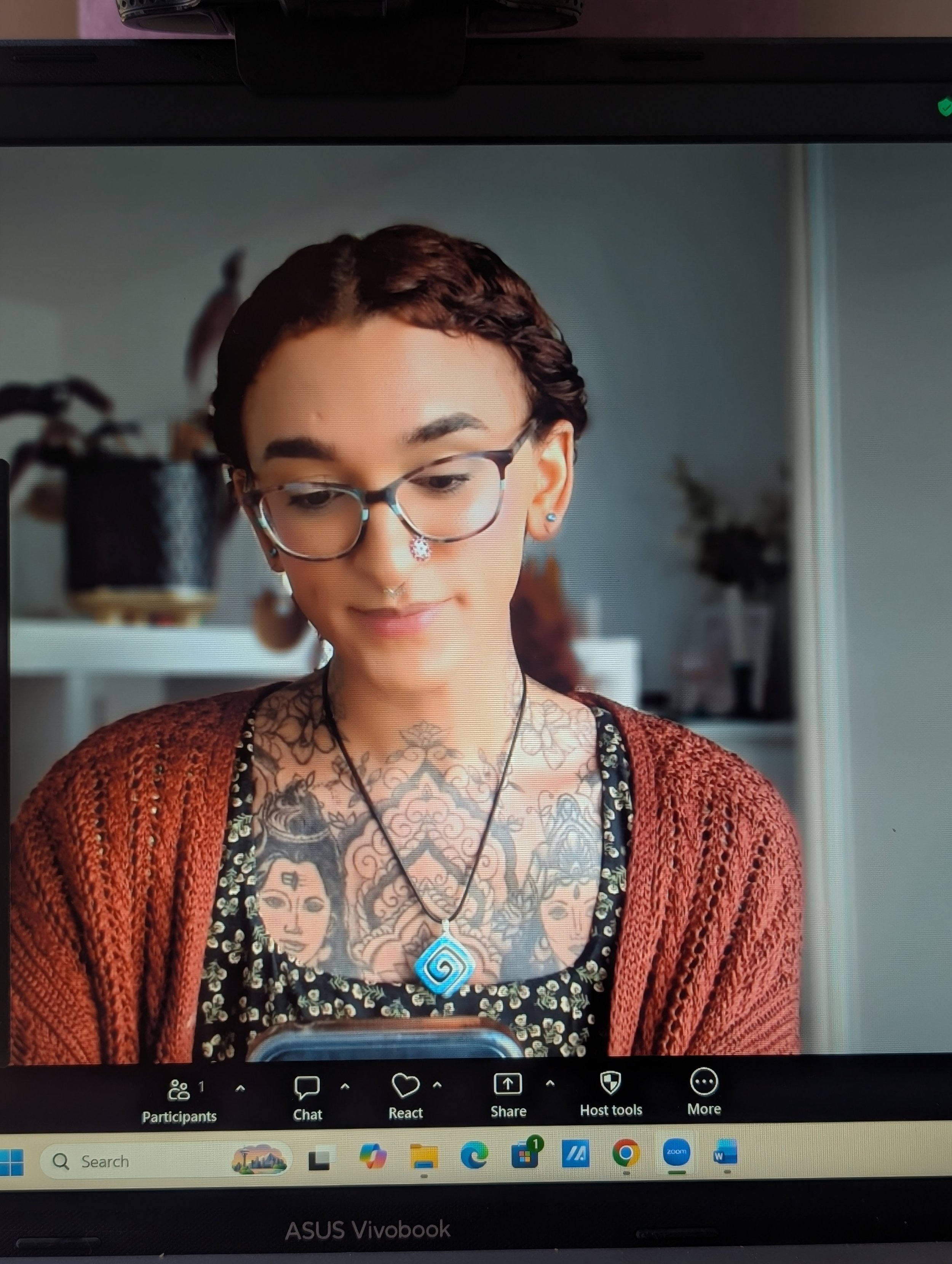 A person with short dark hair and glasses taking a selfie through a computer screen. They are wearing a black top with a floral pattern, a red cardigan, and have numerous tattoos on their chest. They are also wearing a necklace with a blue spiral pendant.