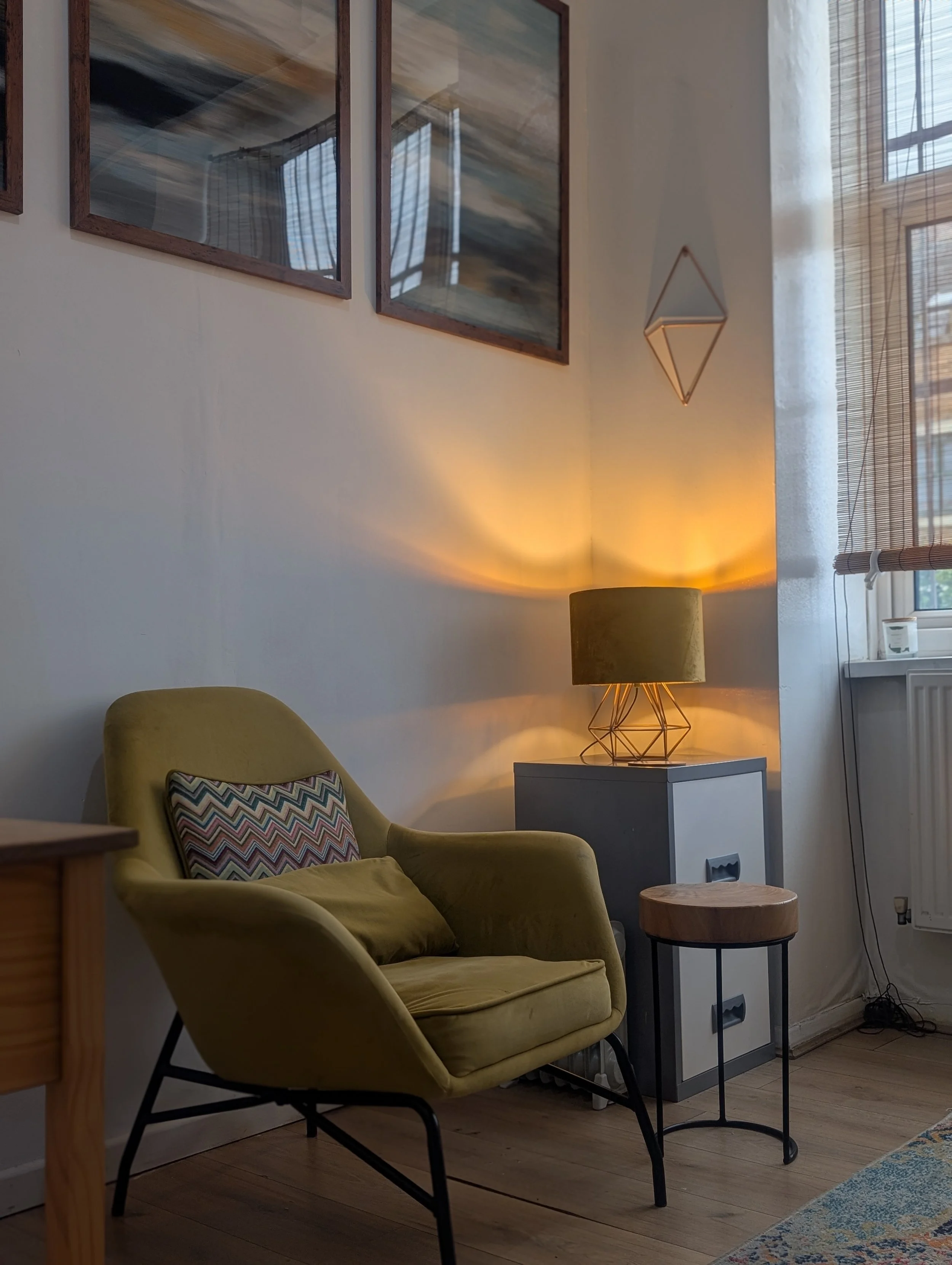 A cozy living room corner with a yellow armchair that has a patterned pillow, a small white side table, a modern geometric table lamp with a beige shade, framed abstract artwork on the wall, a window with bamboo shades, and a colorful rug on a wooden floor.