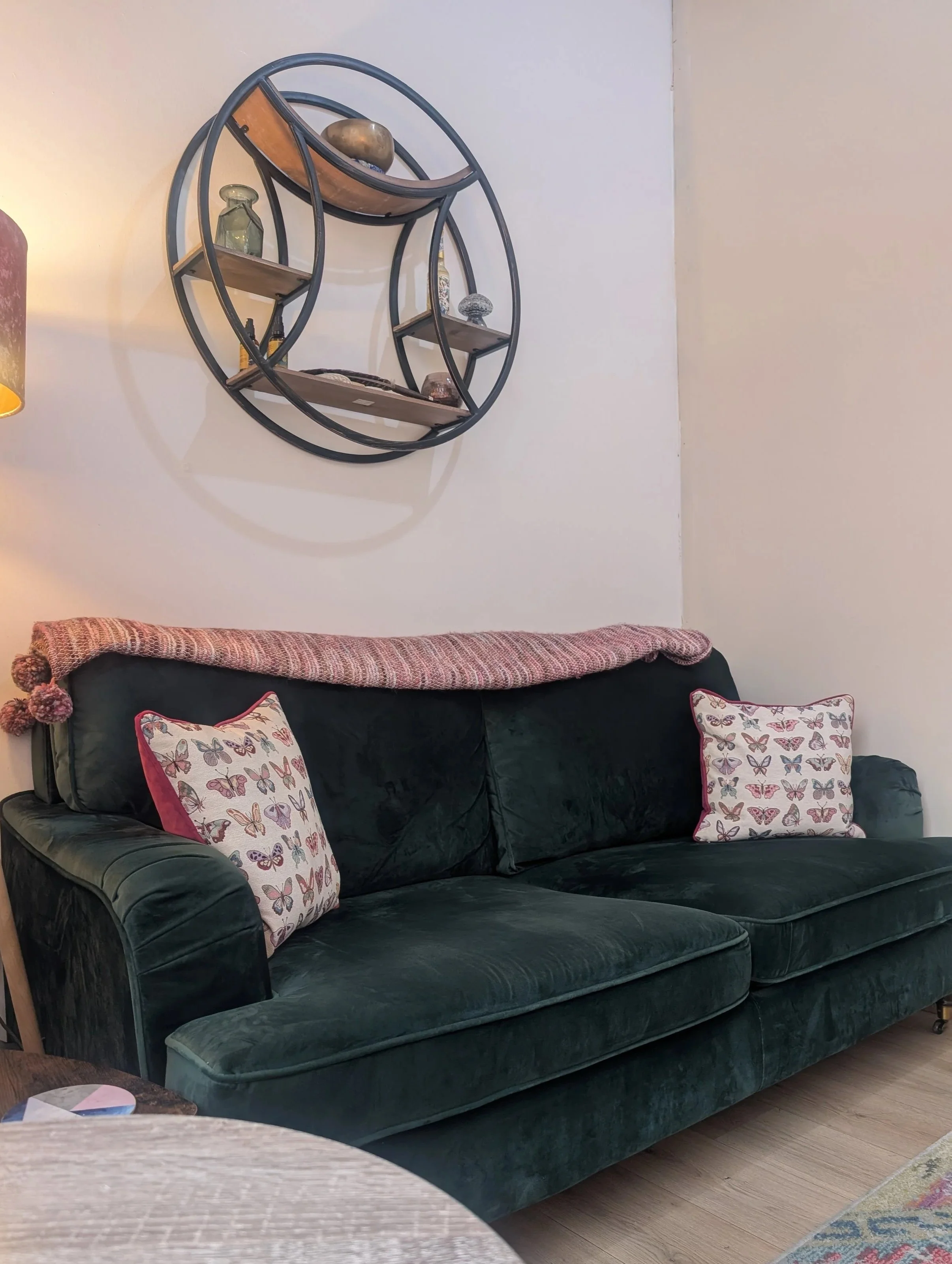 A dark green velvet sofa with two butterfly-patterned pillows and a pink and multicolored blanket draped over the back in a living room with wooden flooring, a side table, and a wall decoration.