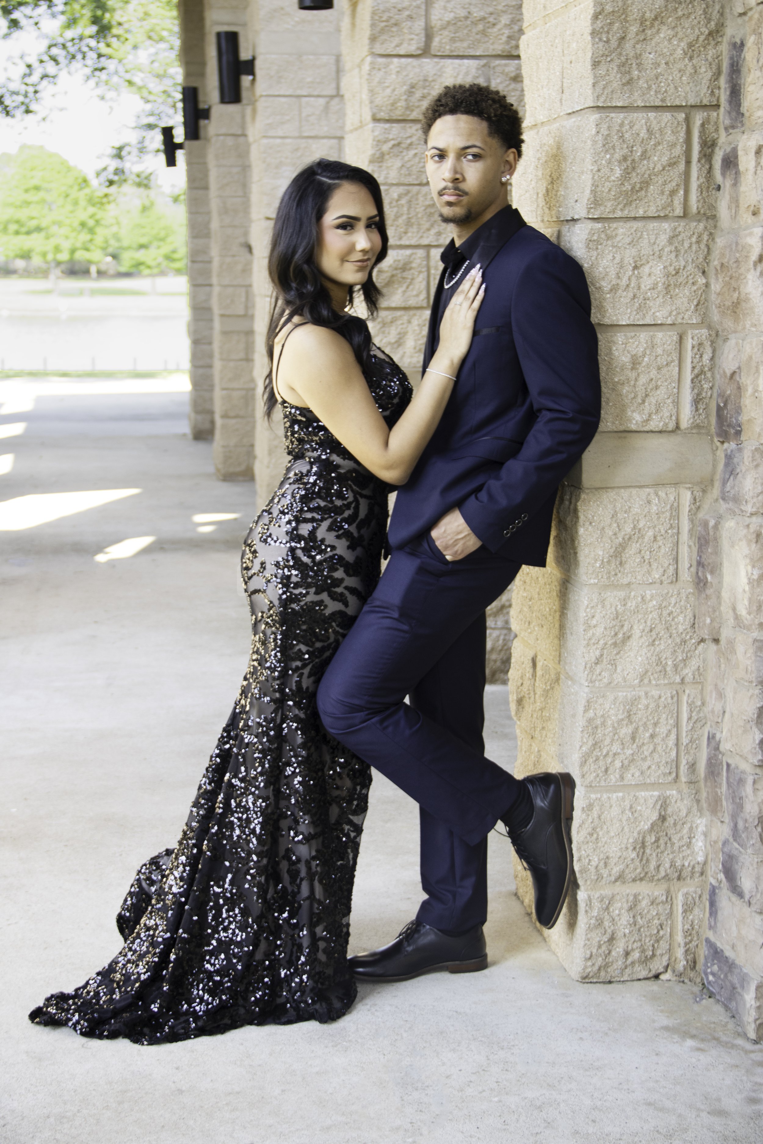 A woman in a black sequin dress and a man in a navy suit pose together in an outdoor setting, leaning against a stone wall.