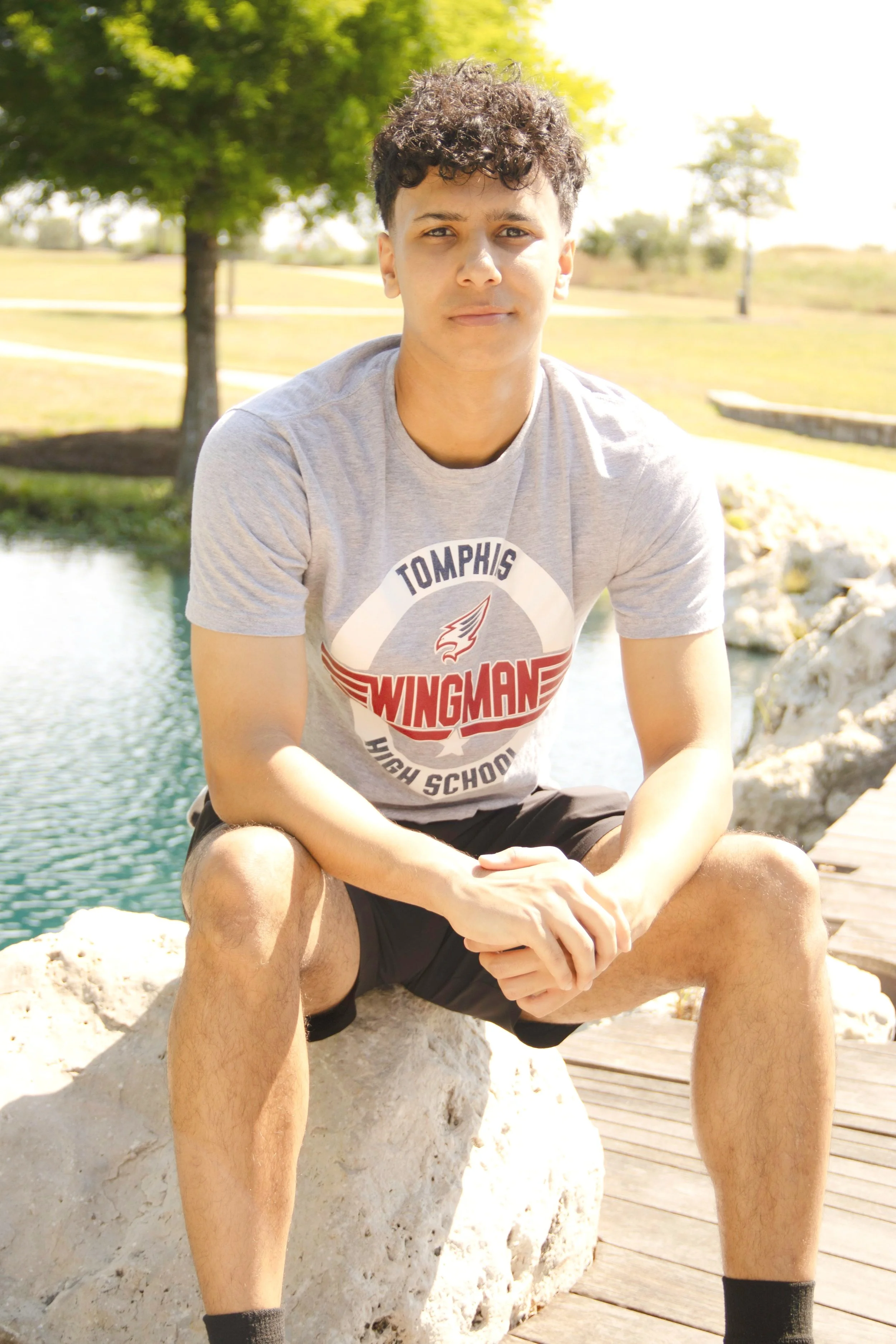 A young man wearing a gray T-shirt with the text 'Tomphis High School' and 'Wingman' printed on it, sitting outdoors on a large rock near a body of water, with trees and a park in the background.