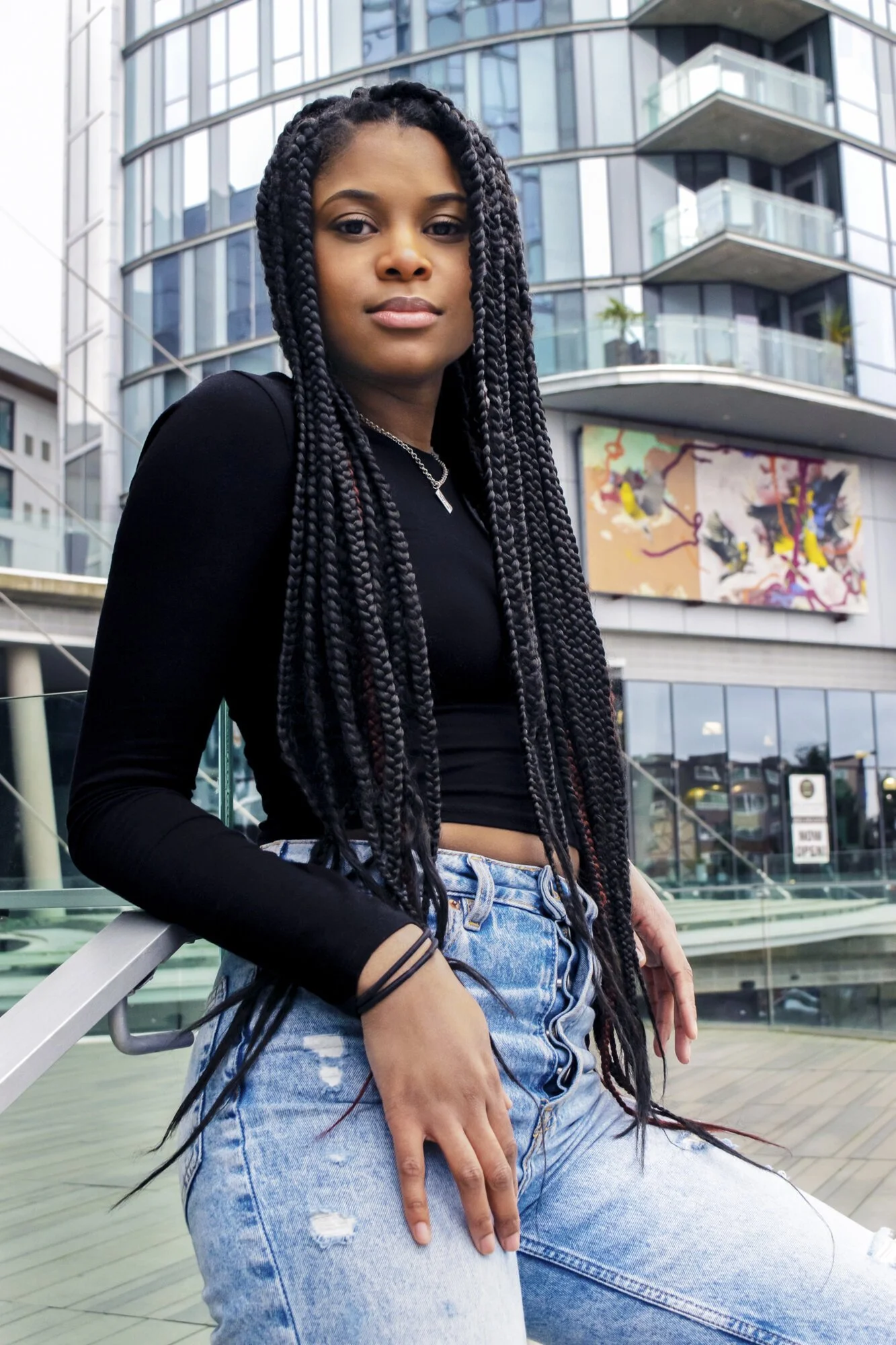 Woman with long braids wearing a black crop top and blue jeans, standing in front of a modern building with glass balconies.