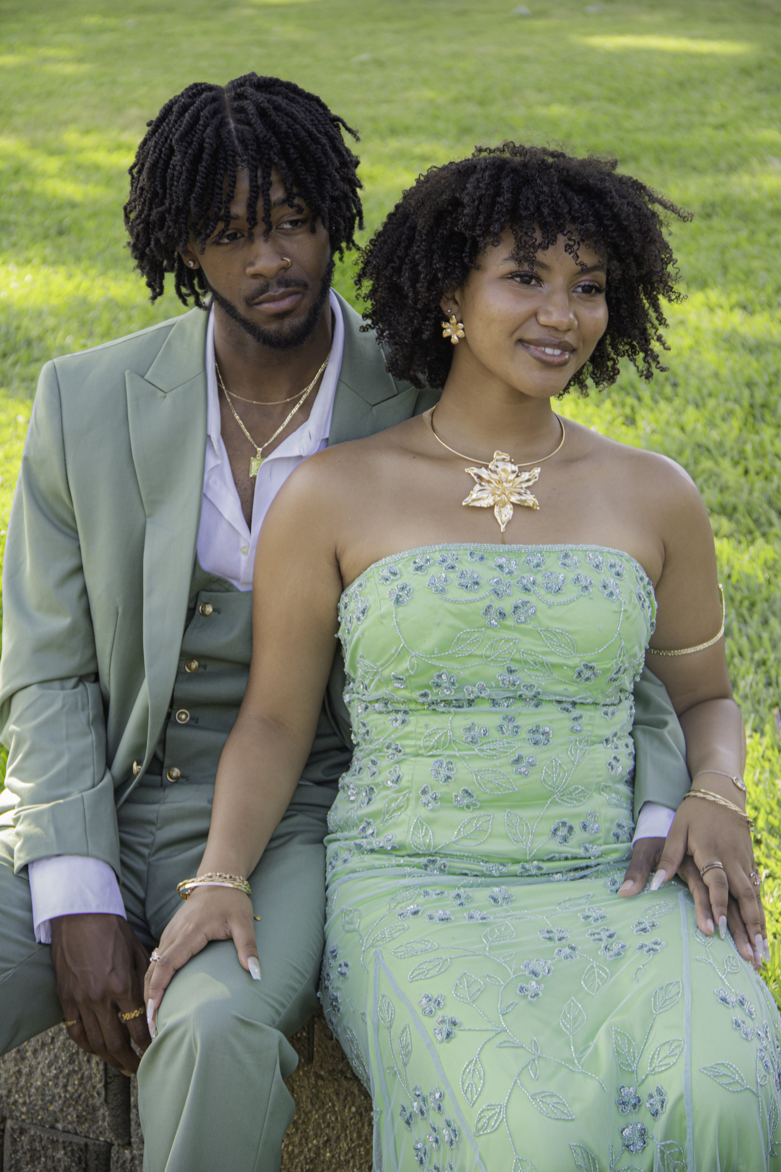 A couple sitting outside on grass. The man wears a light green suit and the woman wears a strapless green dress with floral embroidery. Both have curly hair and are accessorized with gold jewelry.