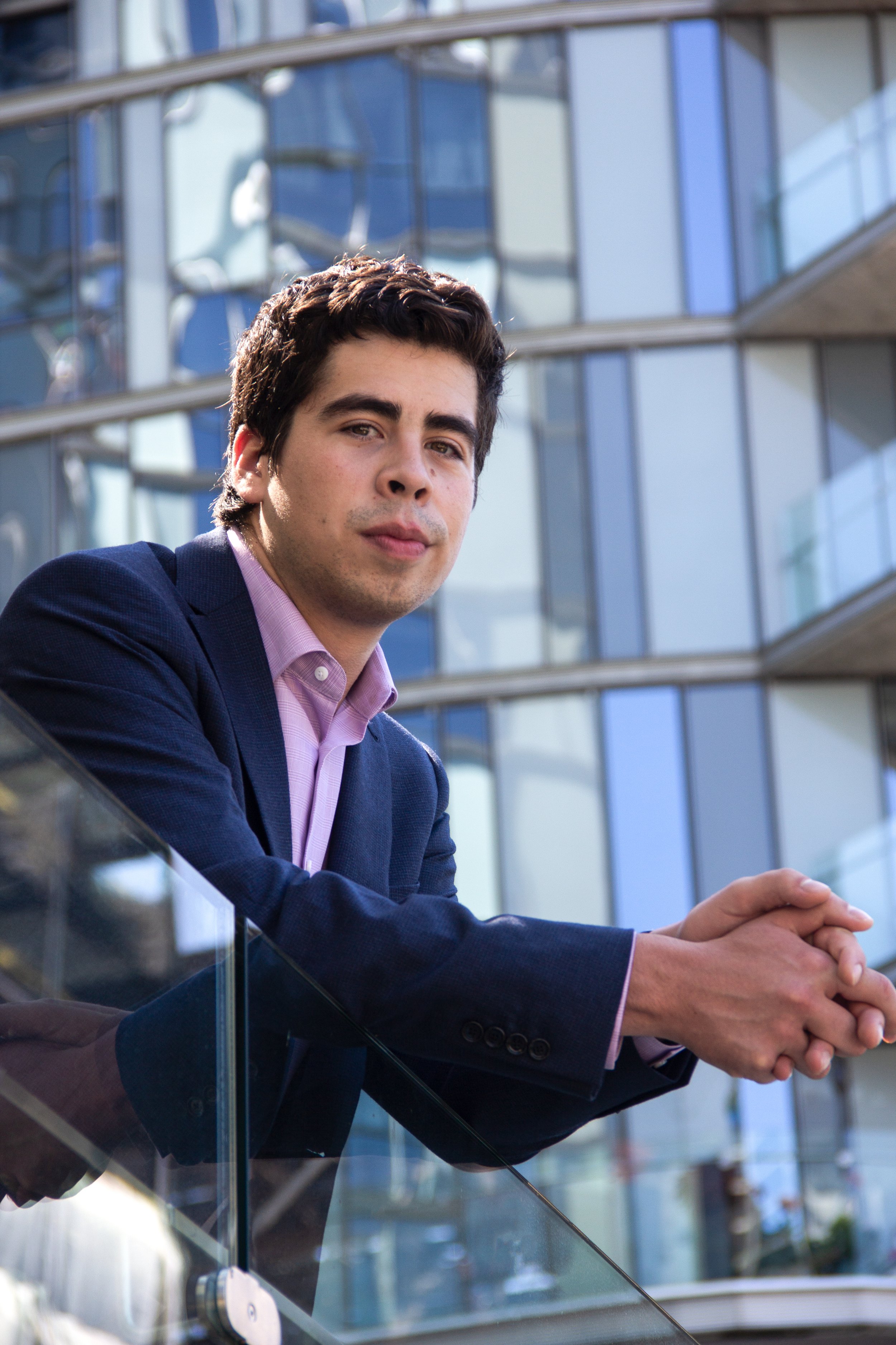 Man in a suit leaning on a glass railing with a modern building in the background.