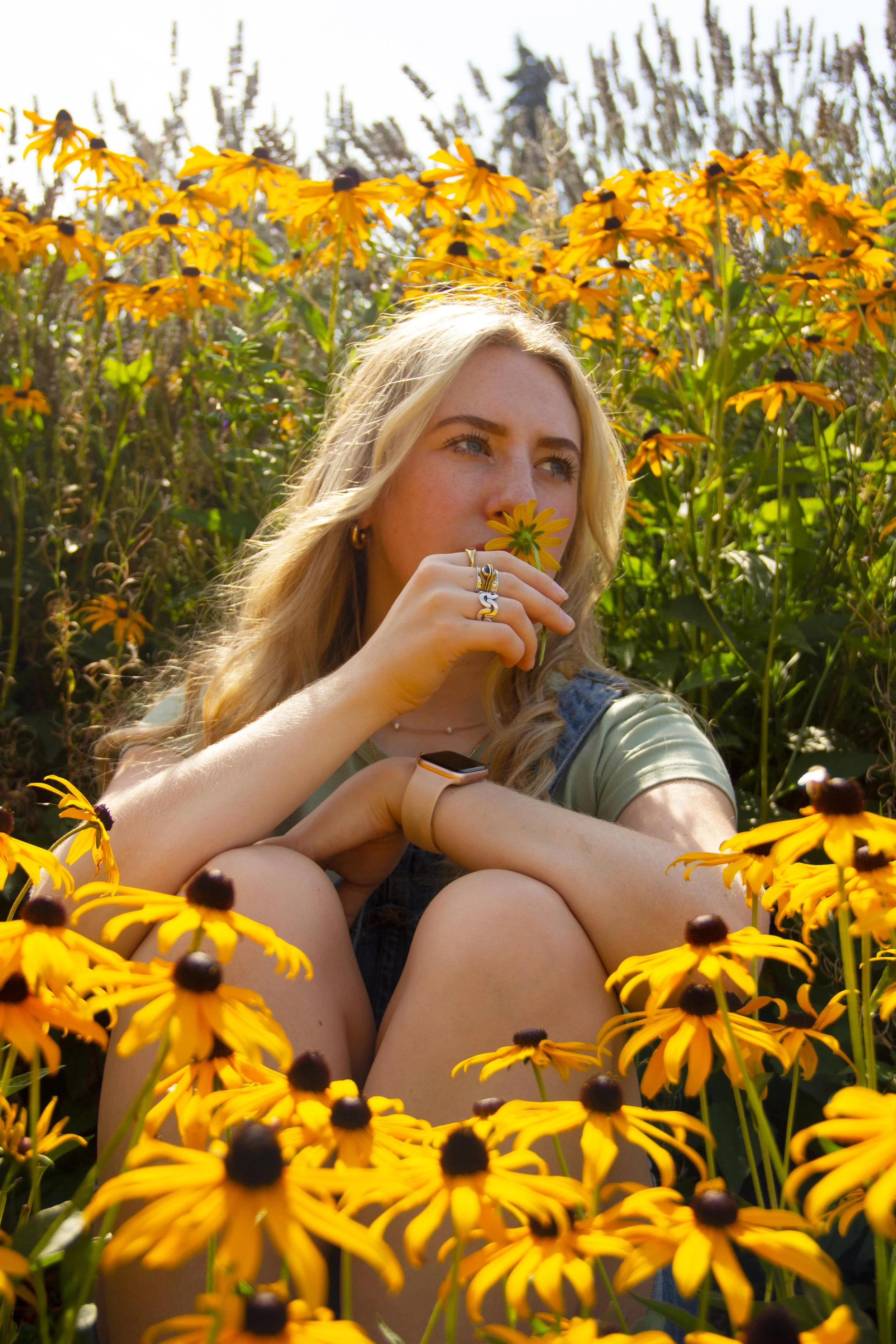 A young woman with blonde hair sitting among yellow flowers holds a flower near her face and looks into the distance