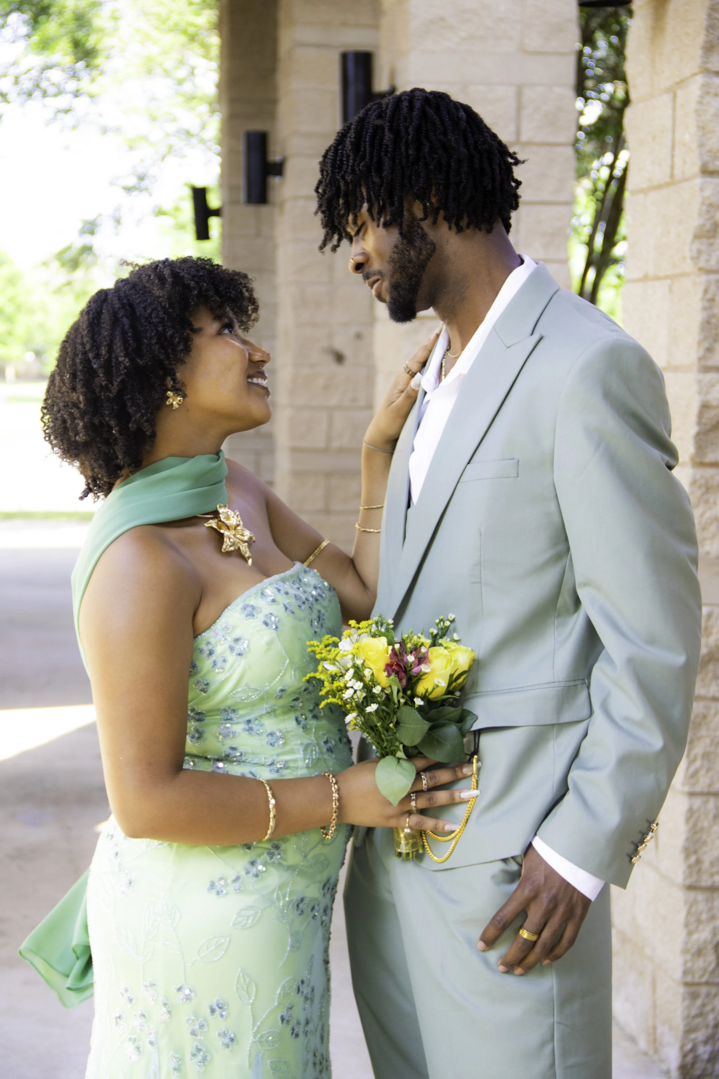 A couple in formal attire, the woman in a light green dress with floral embroidery, holding a bouquet of yellow and purple flowers, talking closely to the man in a matching light green suit.