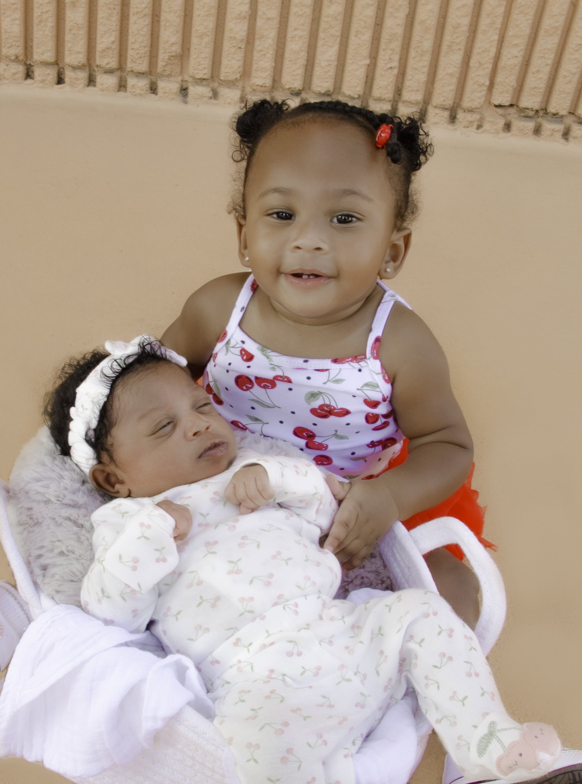 A toddler in a cherry-patterned outfit is sitting next to a newborn baby in a floral onesie. The baby is asleep in a basket lined with a soft blanket. The toddler has a small clip in their hair.