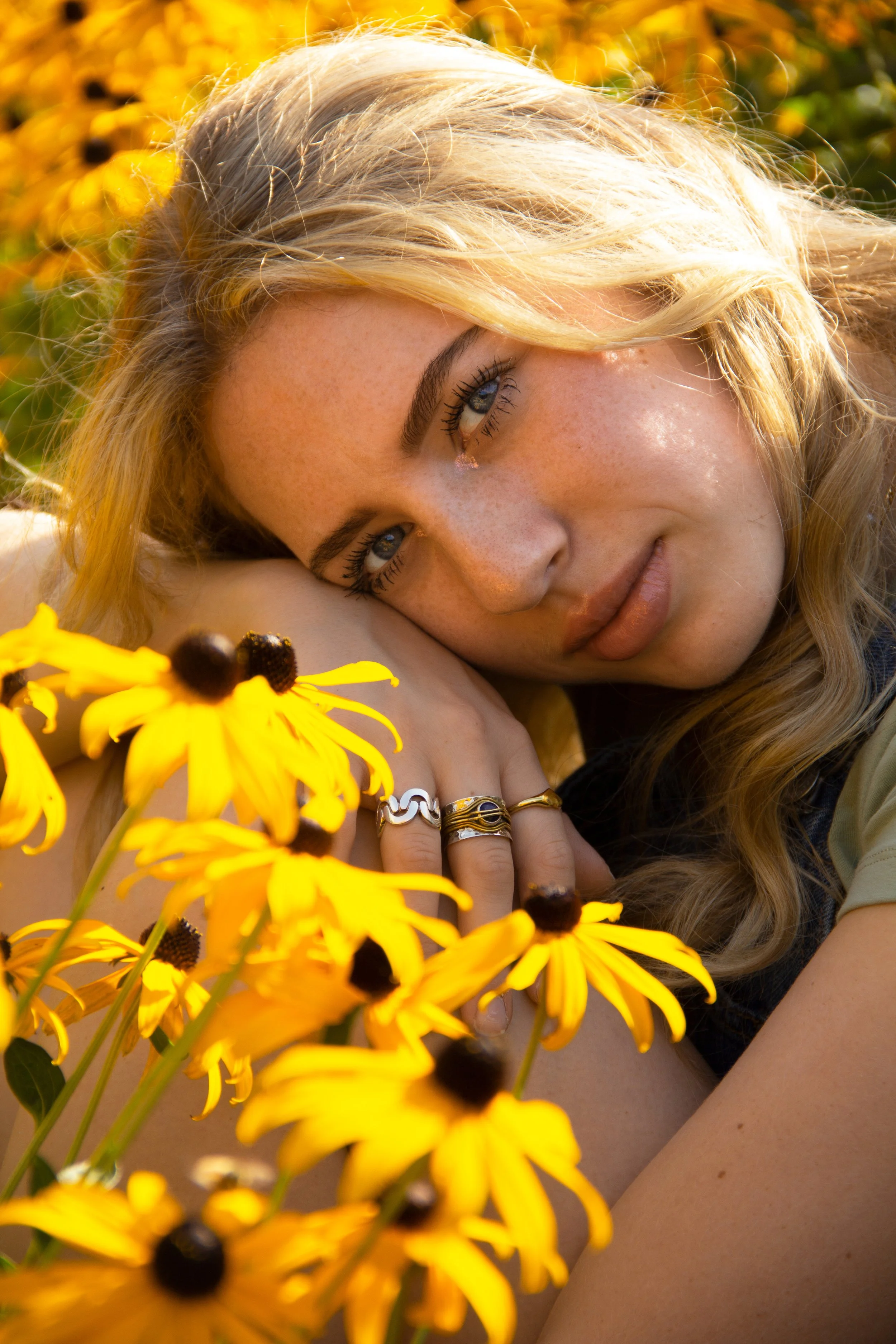 Person with long blonde hair lying among yellow flowers, wearing silver and gold rings.