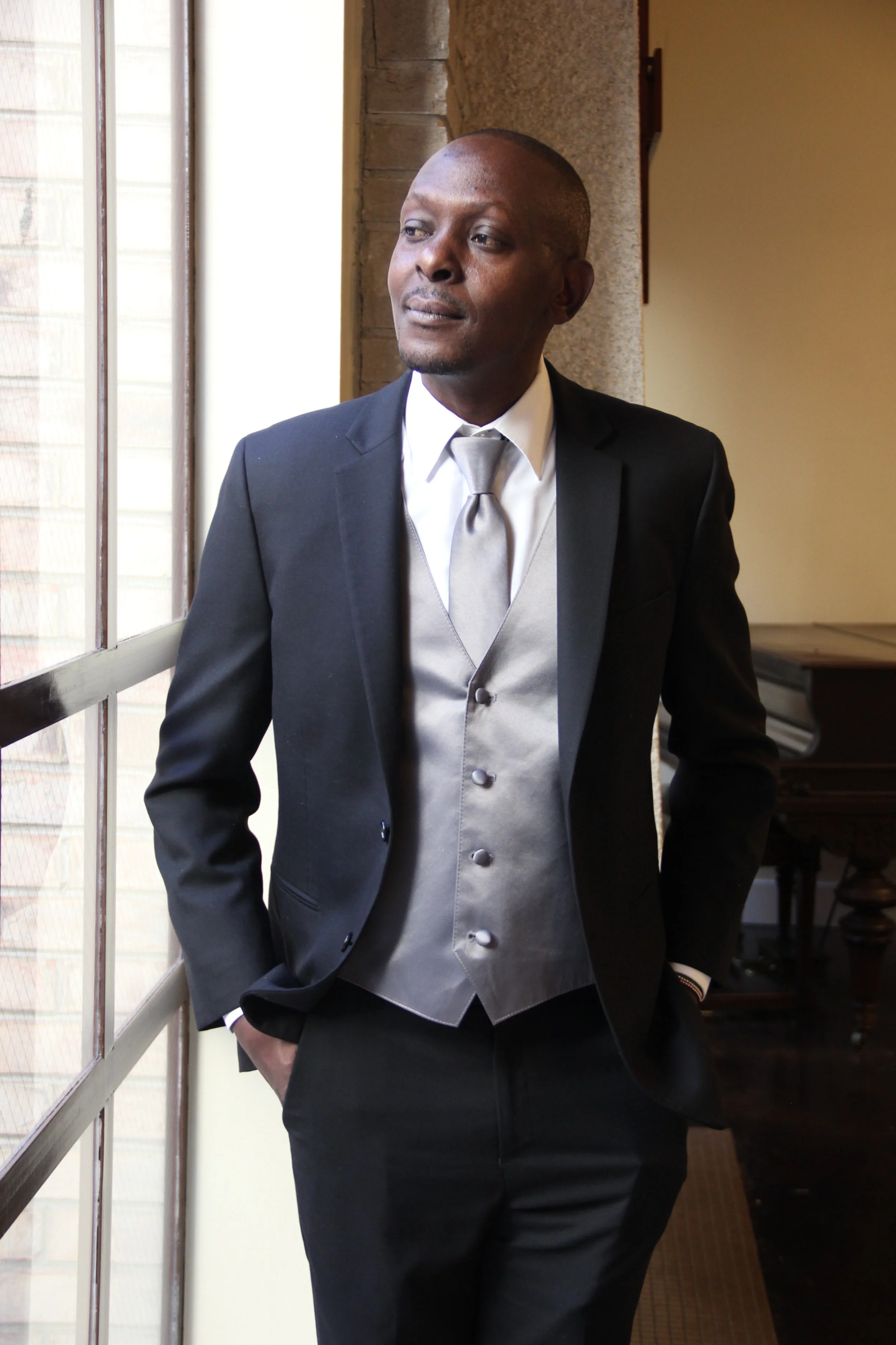 A man in a suit with a gray vest and tie, standing by a window looking outside.