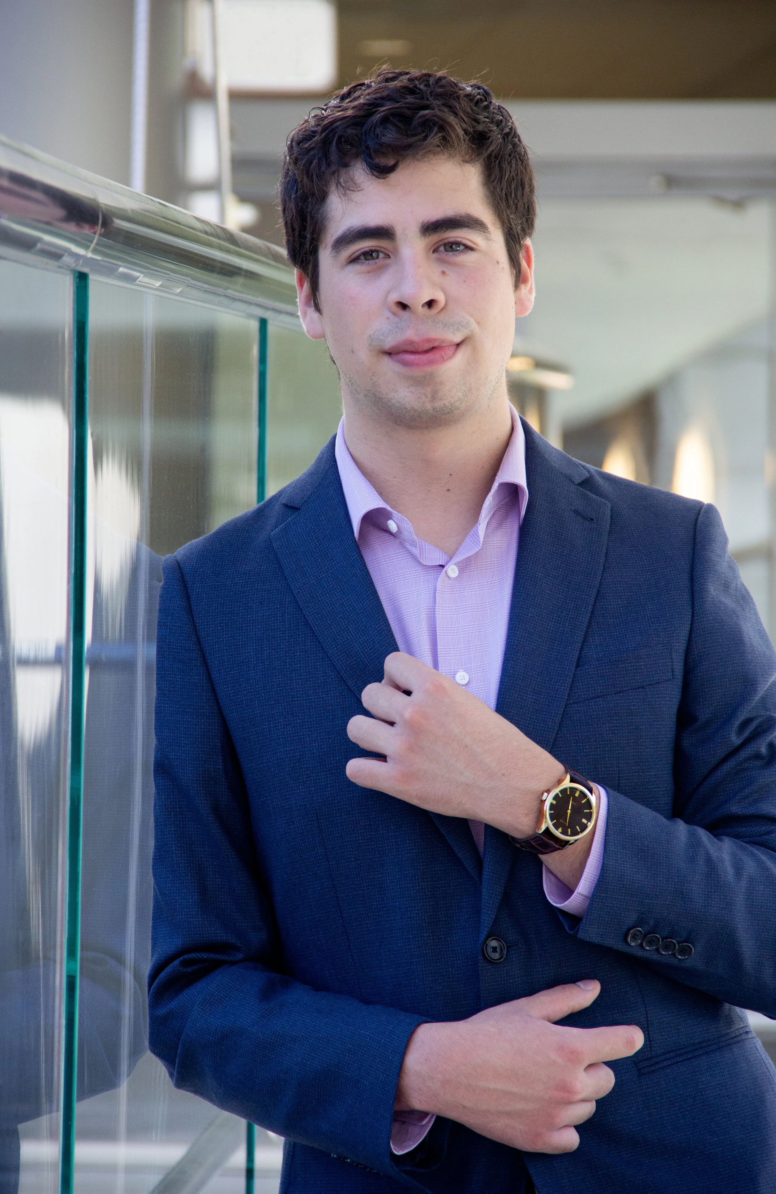 Young man wearing a dark blue suit with a light purple shirt, standing outdoors near glass panels, adjusting his jacket and wearing a watch.