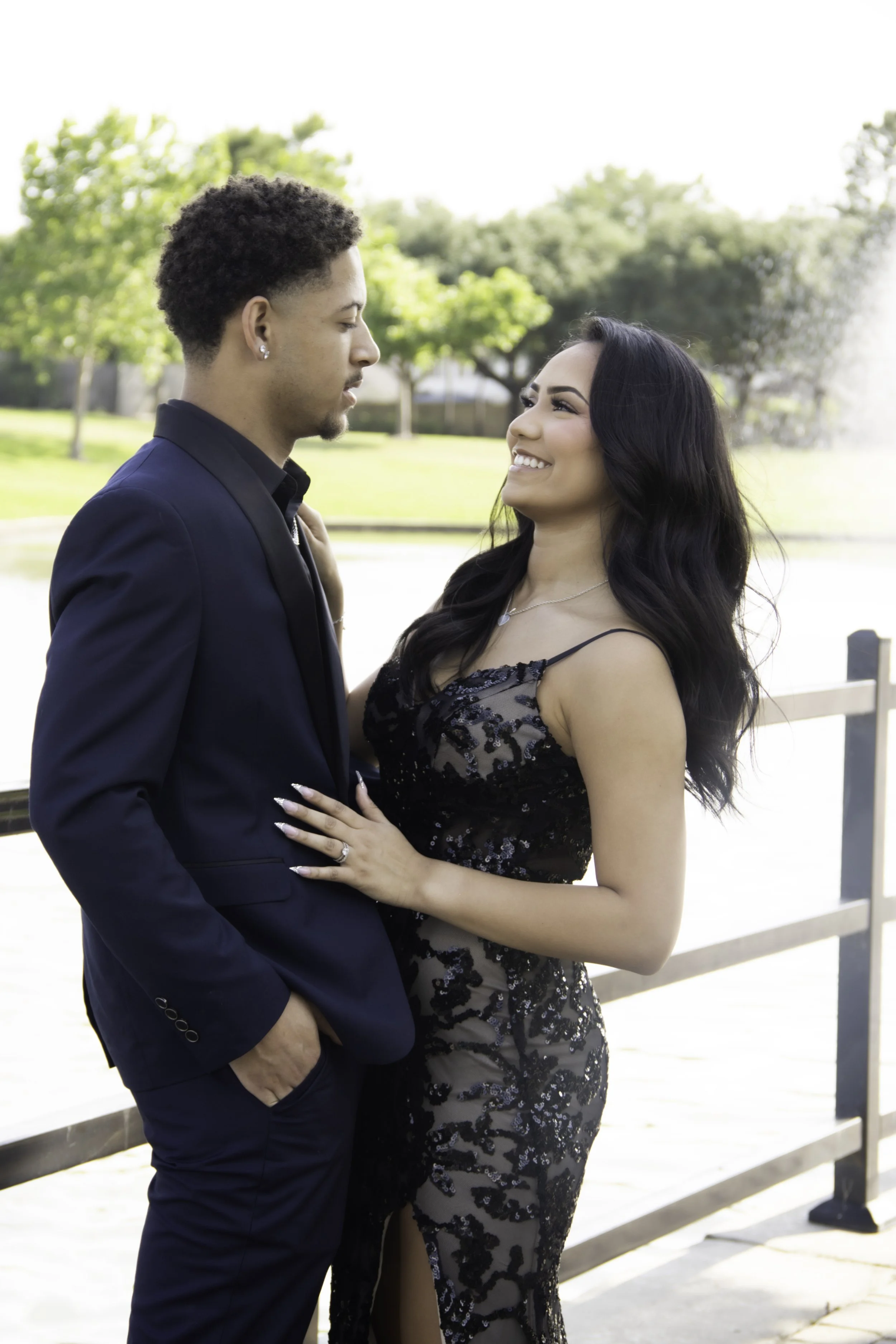 A couple dressed in formal attire stands by a railing near a pond, with trees and a fountain in the background. The woman is smiling while looking at the man.