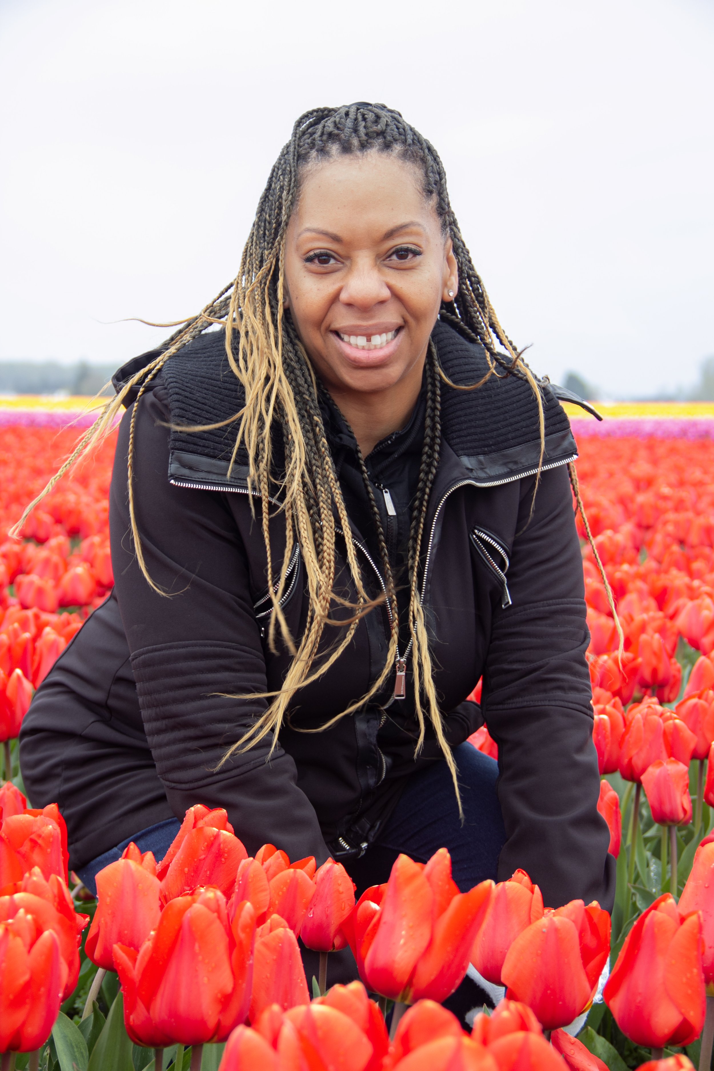 Person posing in a field of red tulips with braided hair and a black jacket, smiling towards the camera.
