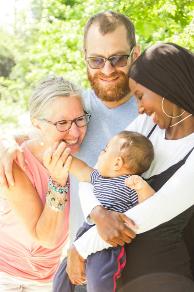 A happy family outdoors, with two adults smiling at a baby held by a third adult, surrounded by green foliage.