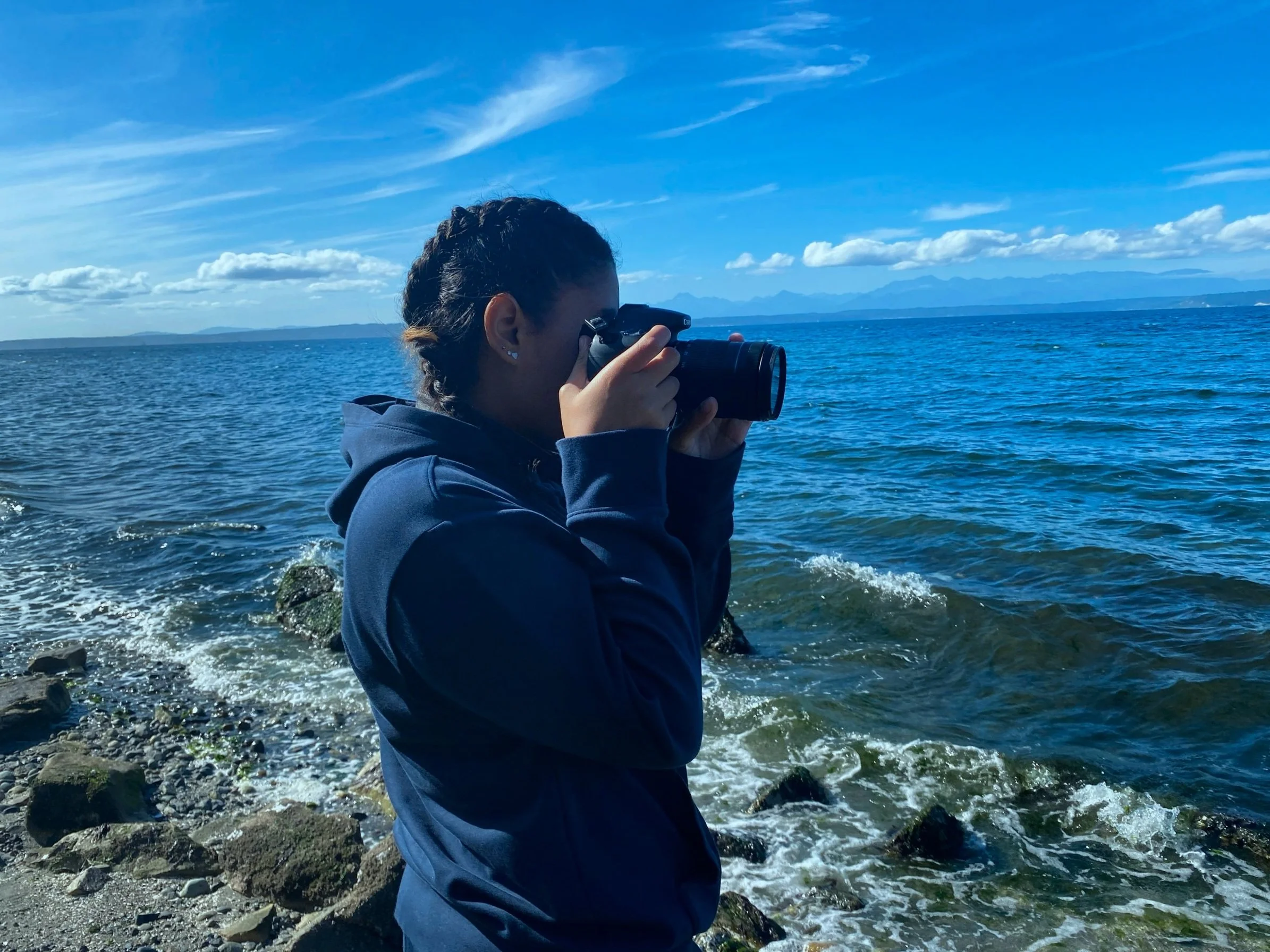 Person in a navy jacket taking a photo with a camera by the ocean, blue sky with clouds, mountains in the distance.