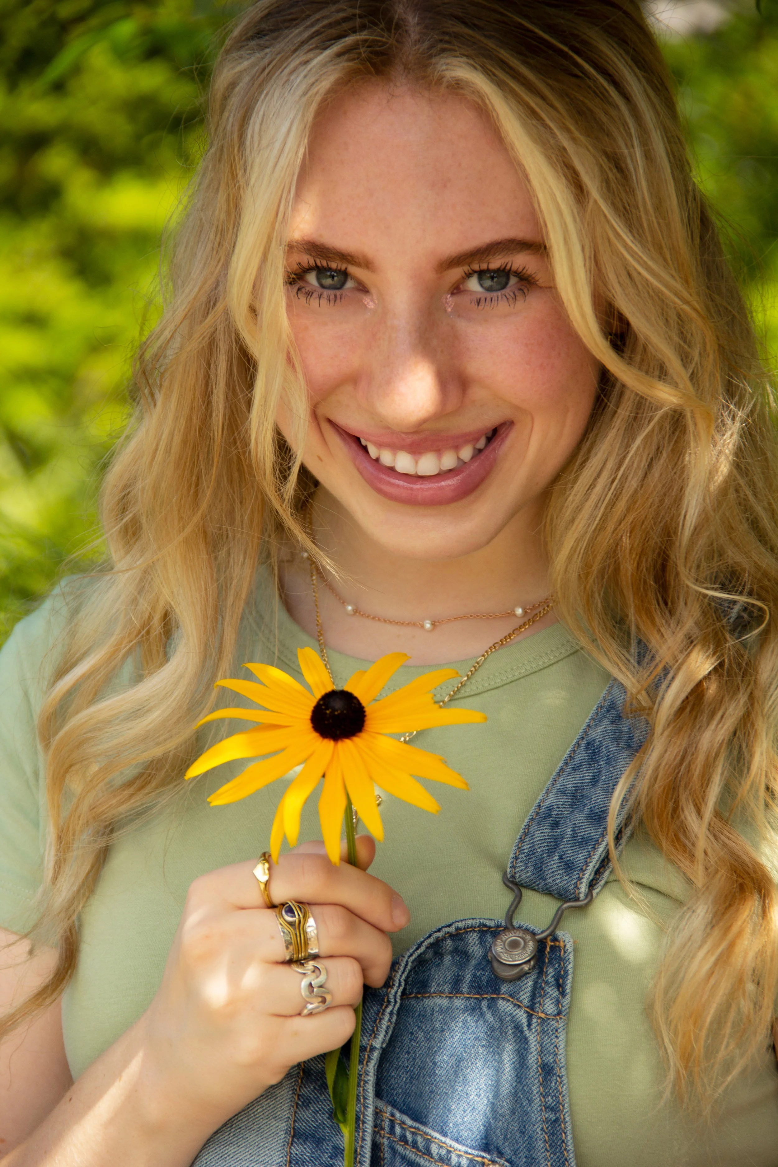 A smiling person with long blonde hair, wearing a green top and denim overalls, holding a yellow flower with a dark center.