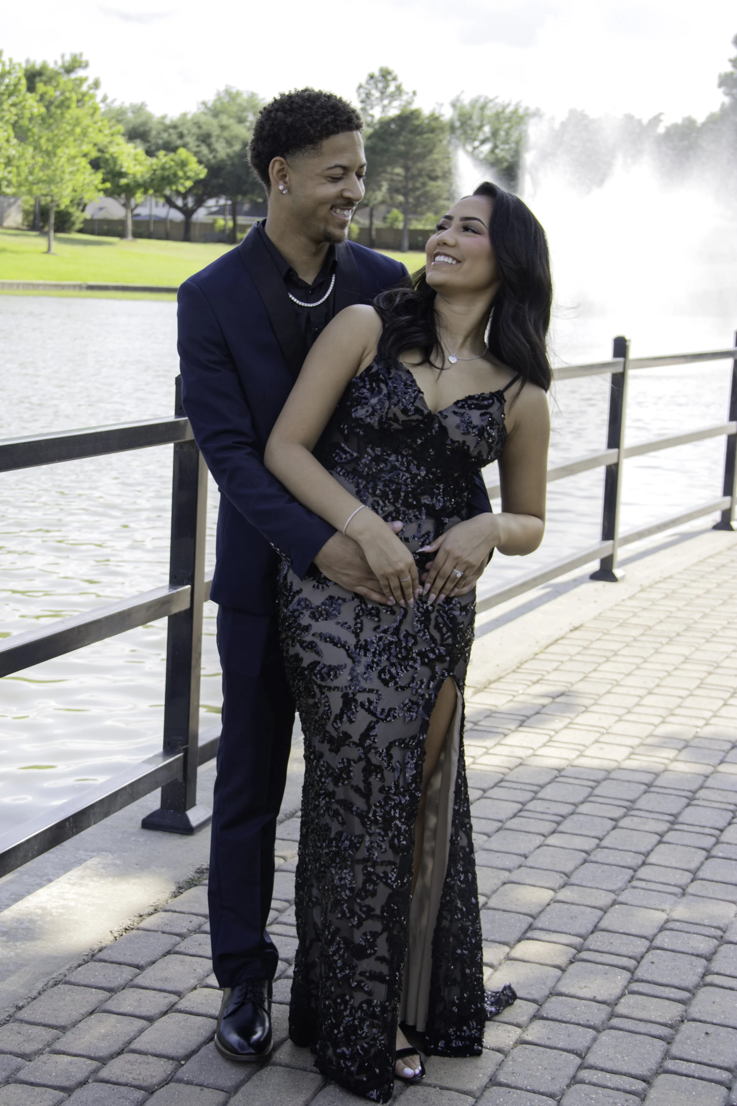 A couple smiling while embracing near a lake, with a fountain in the background. They are dressed in formal attire, standing on a paved walkway next to a railing.