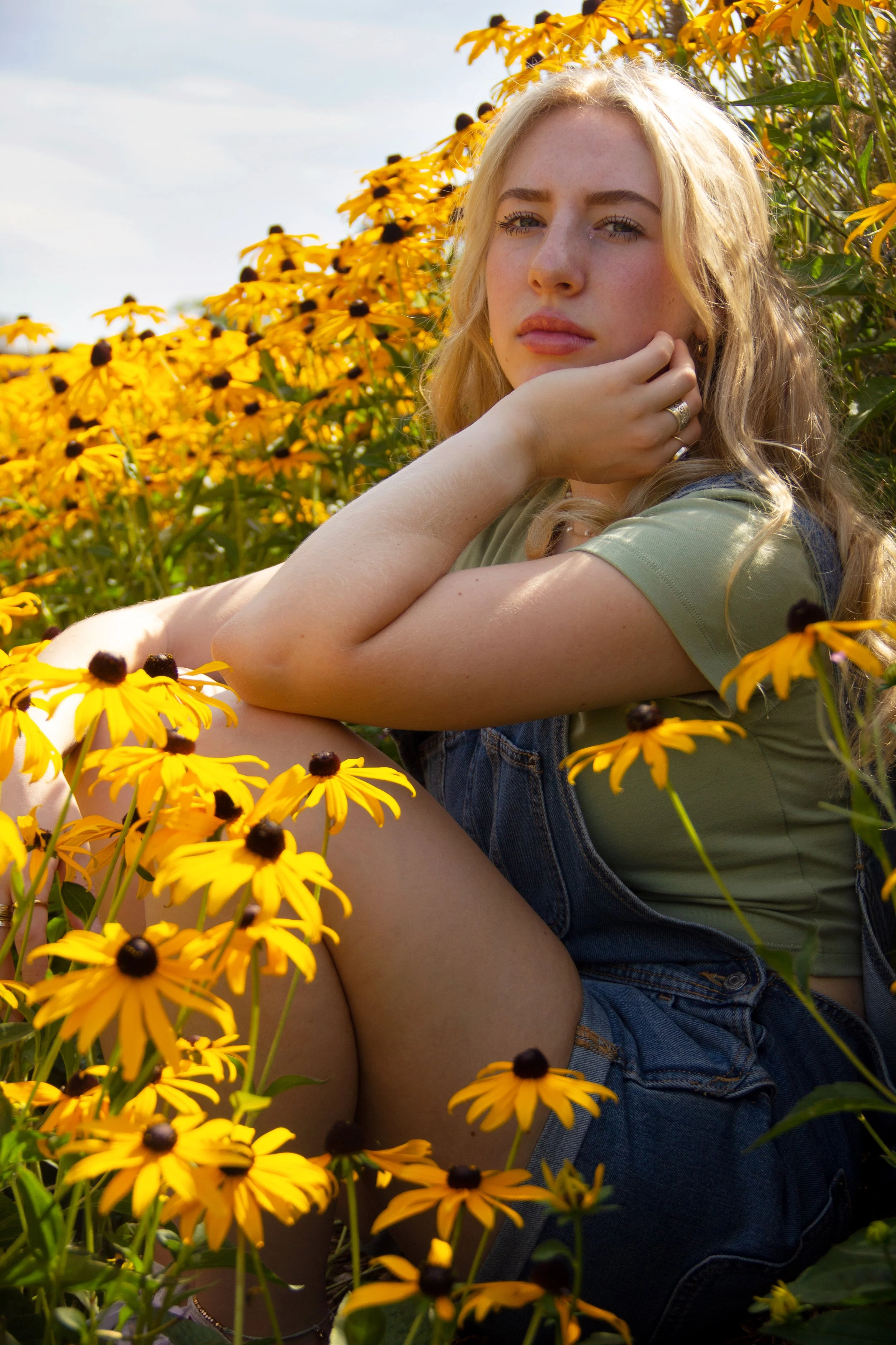 A young woman with long blonde hair sitting among yellow flowers in a field, wearing a green shirt and denim overalls, looking contemplative.